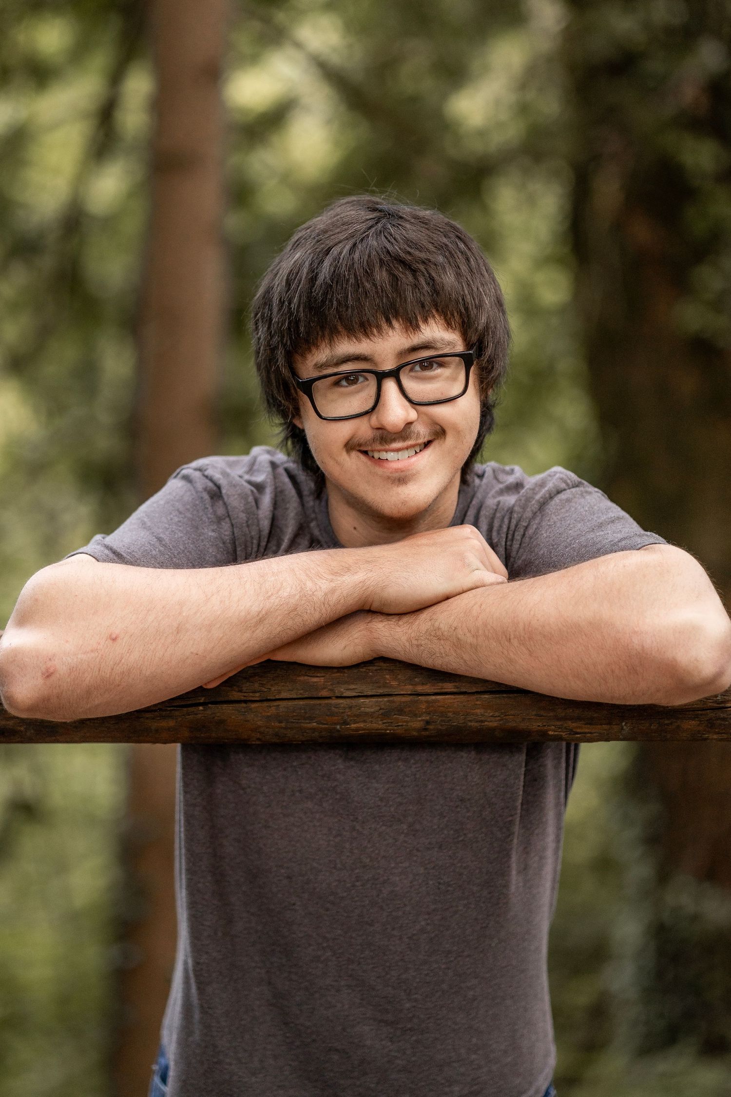 Person in glasses and grey shirt leans on wooden railing in forest setting.