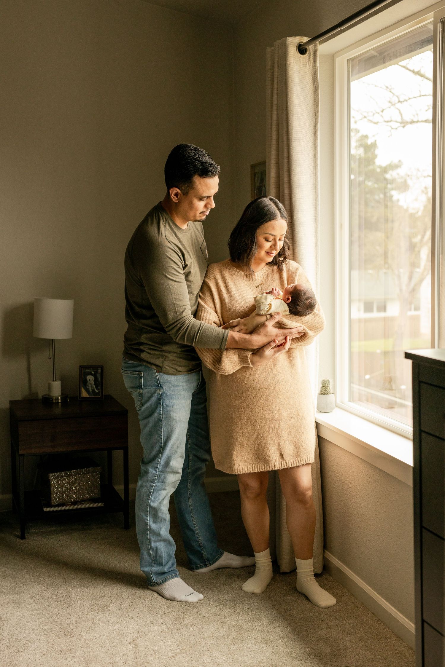 A couple stands by a sunny window in a cozy bedroom holding their newborn baby in soft, natural light.