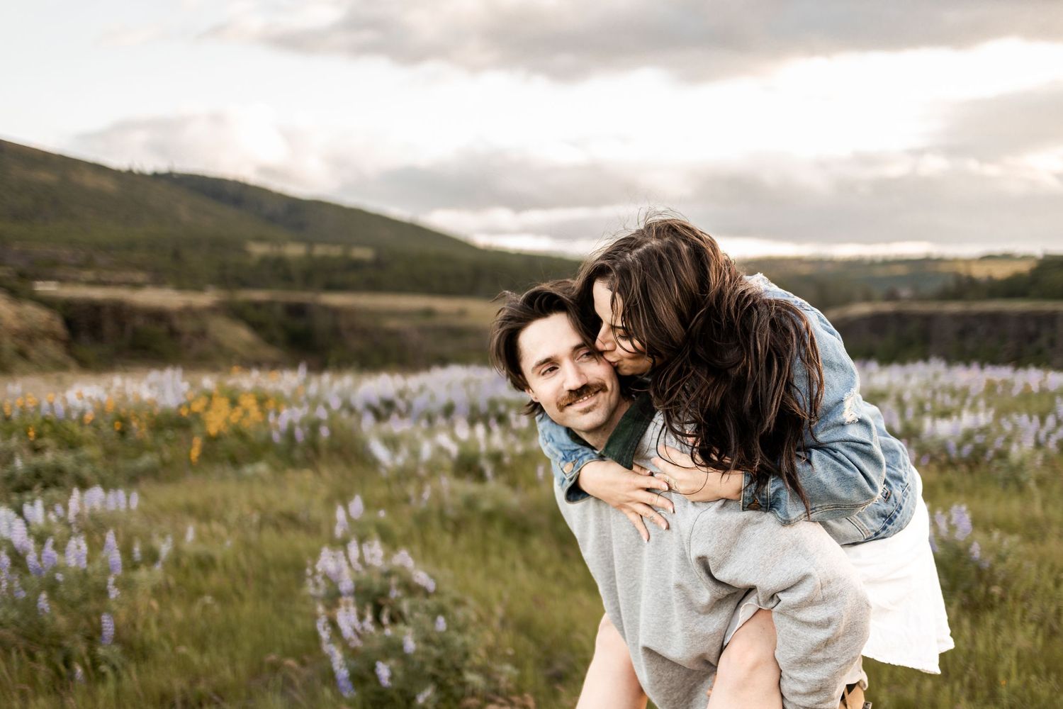 A happy couple in a flower field, woman kissing man on cheek while he gives her a piggyback ride.