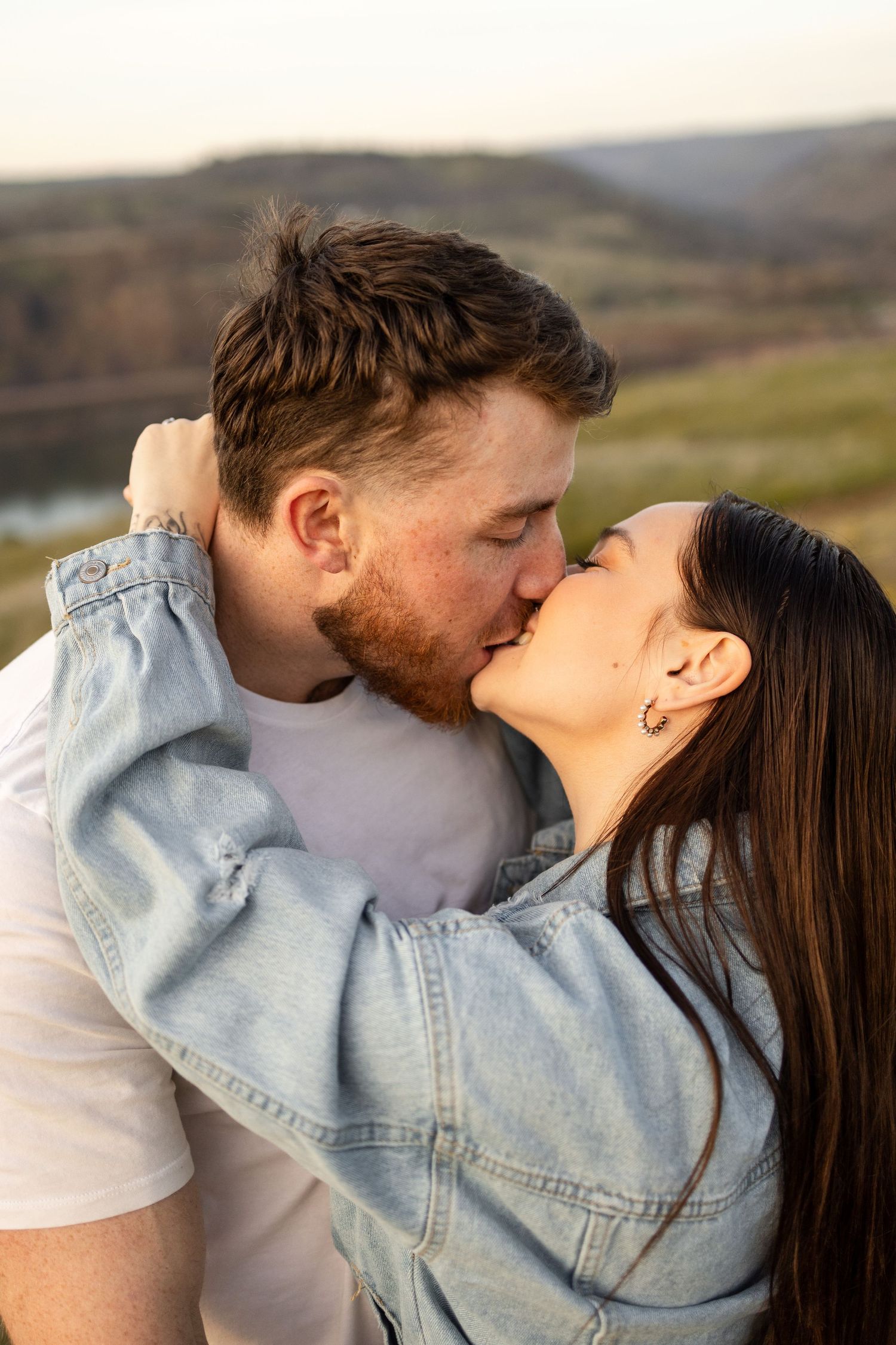 Couple sharing an intimate kiss in denim clothing against a scenic mountain landscape backdrop at sunset. Columbia River Gorge.