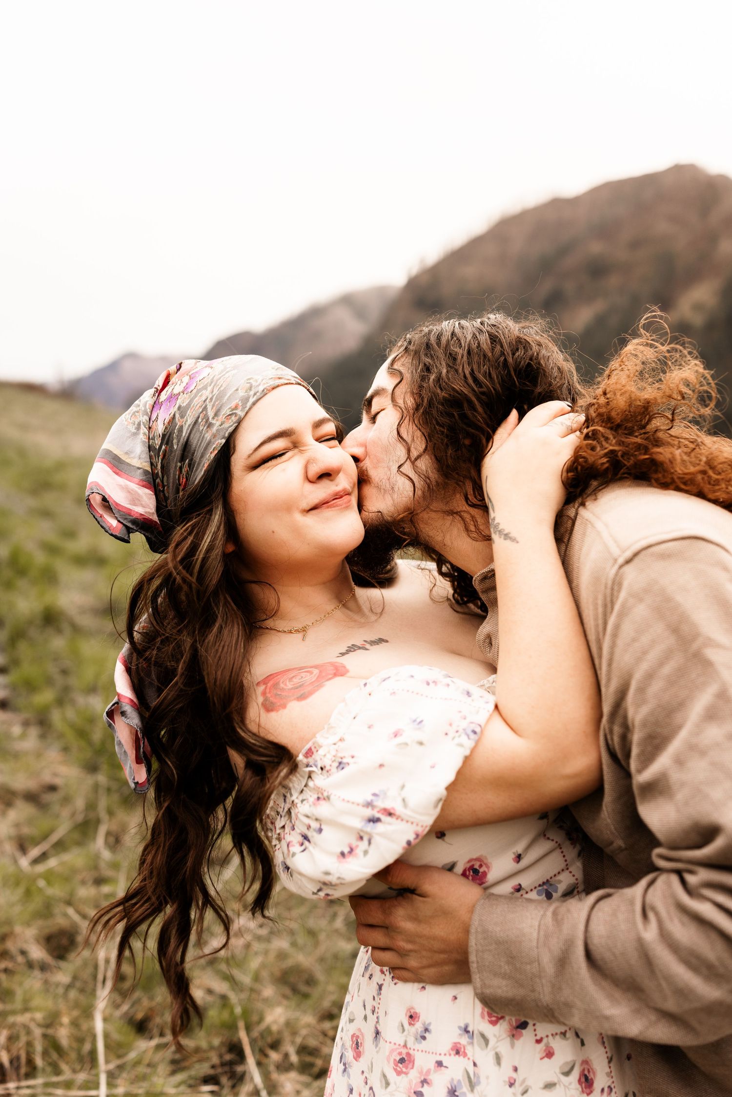 A couple shares a romantic kiss outdoors with mountains in the background, woman wearing floral dress and headscarf.