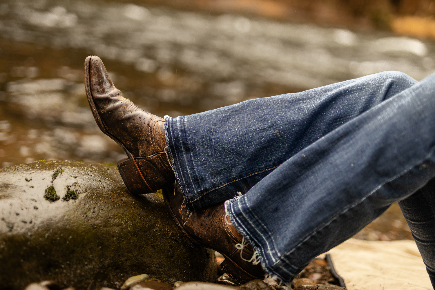 Person relaxing outdoors wearing blue jeans and brown cowboy boots resting on a rock near a stream.