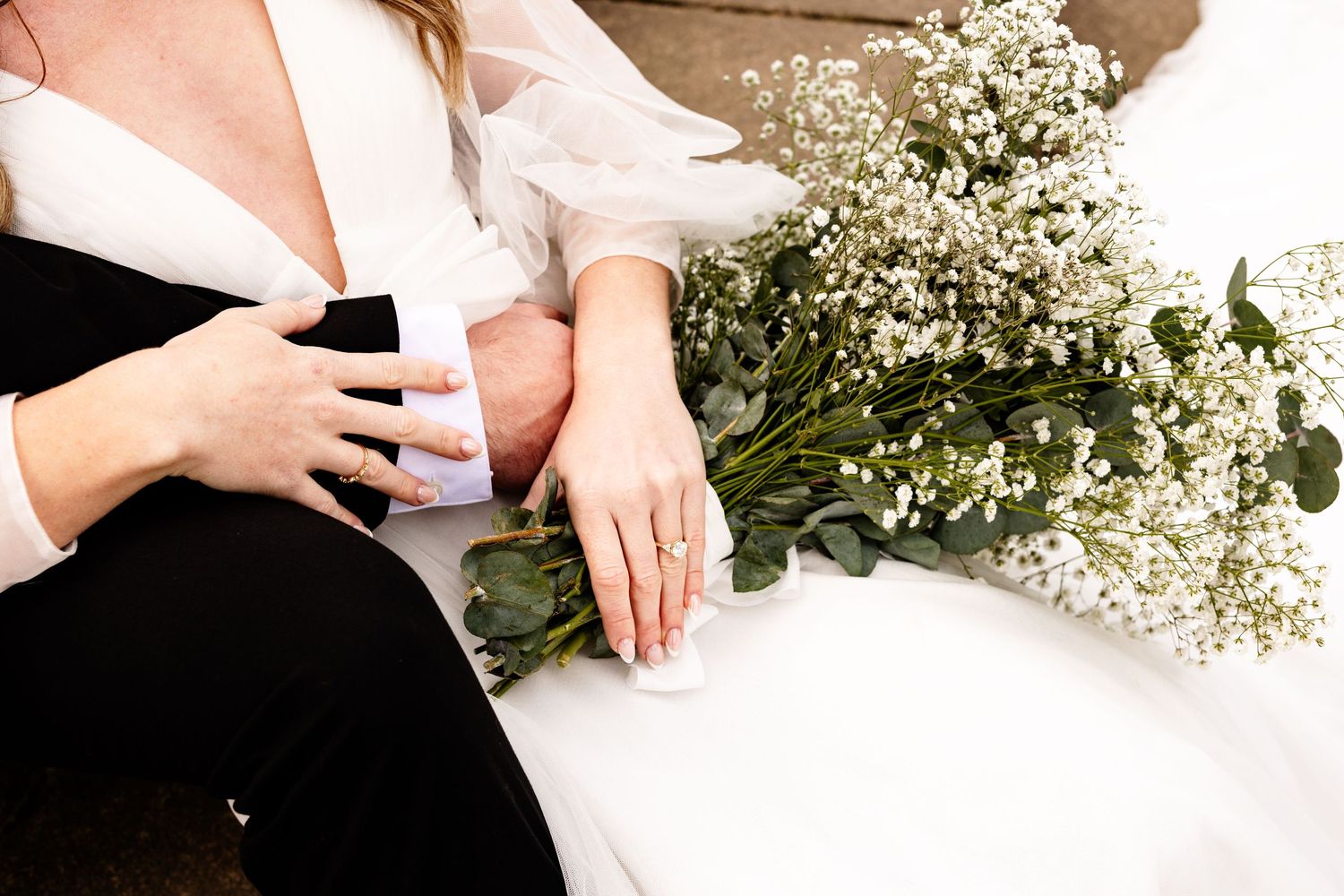 Bride and bridesmaid sitting together holding a white bouquet of baby's breath and greenery, wearing rings.