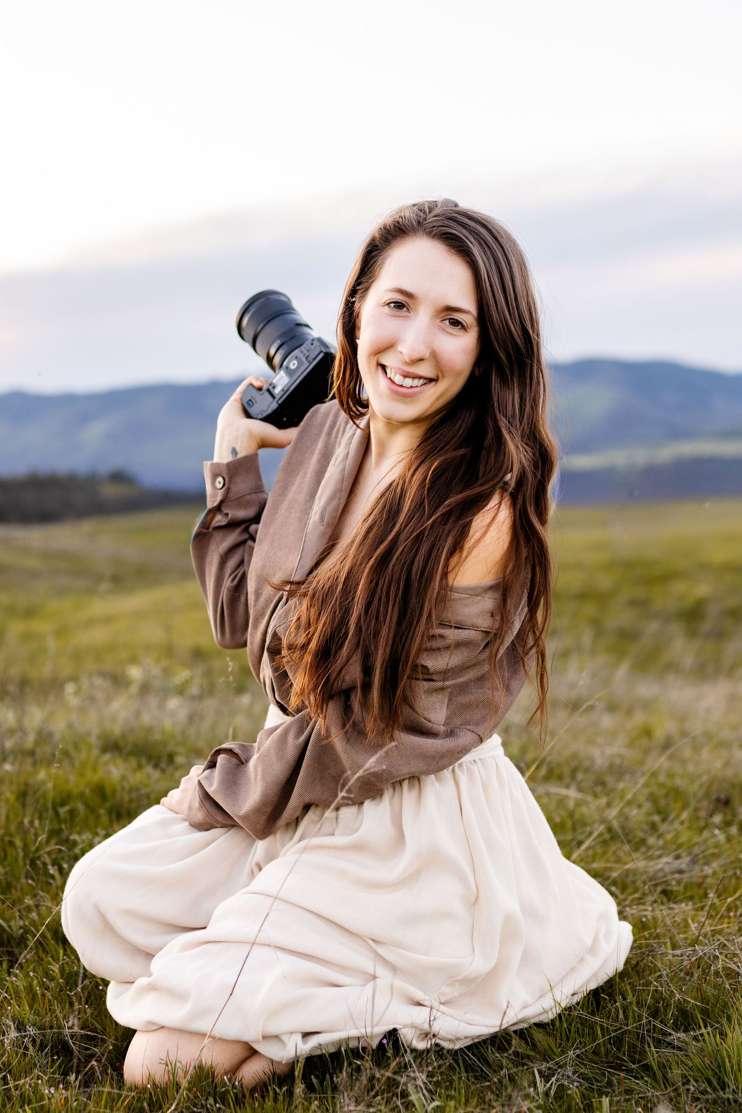 Smiling woman with long brown hair holding a camera, sitting in a green meadow with mountains in the background.