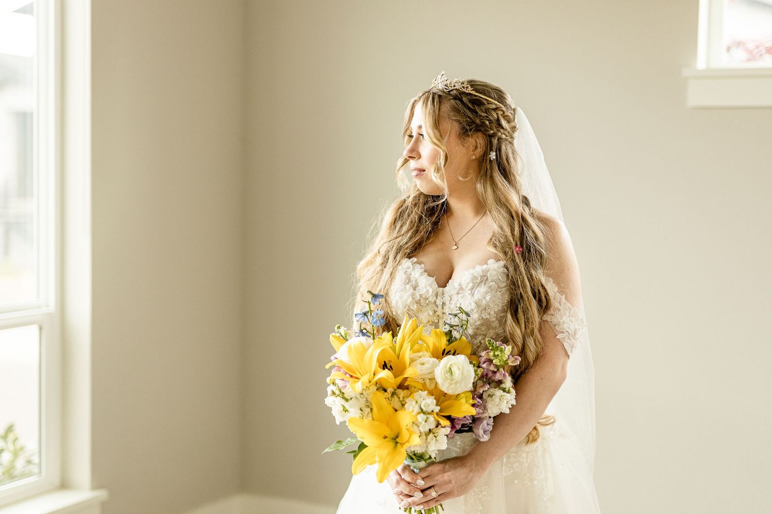 Bride in lace wedding dress holding vibrant yellow floral bouquet, standing by window in soft natural light.