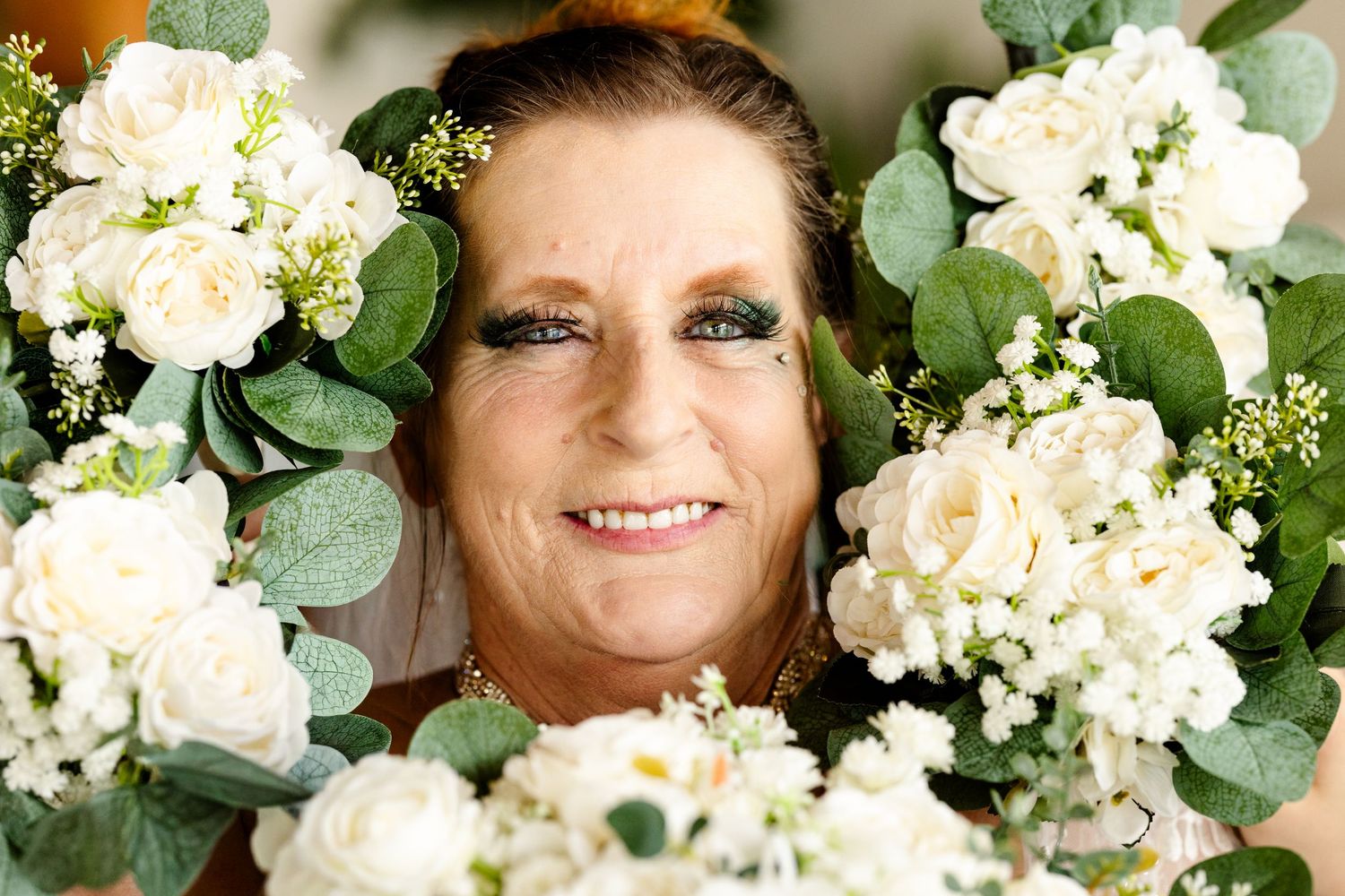 Smiling woman surrounded by lush white roses and eucalyptus greenery in a beautiful floral arrangement portrait.