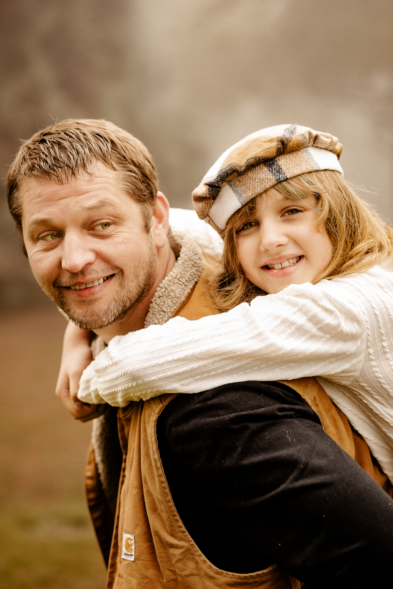 A smiling man gives a happy young girl a piggyback ride outdoors, both wearing warm casual clothing in a cozy autumn setting.