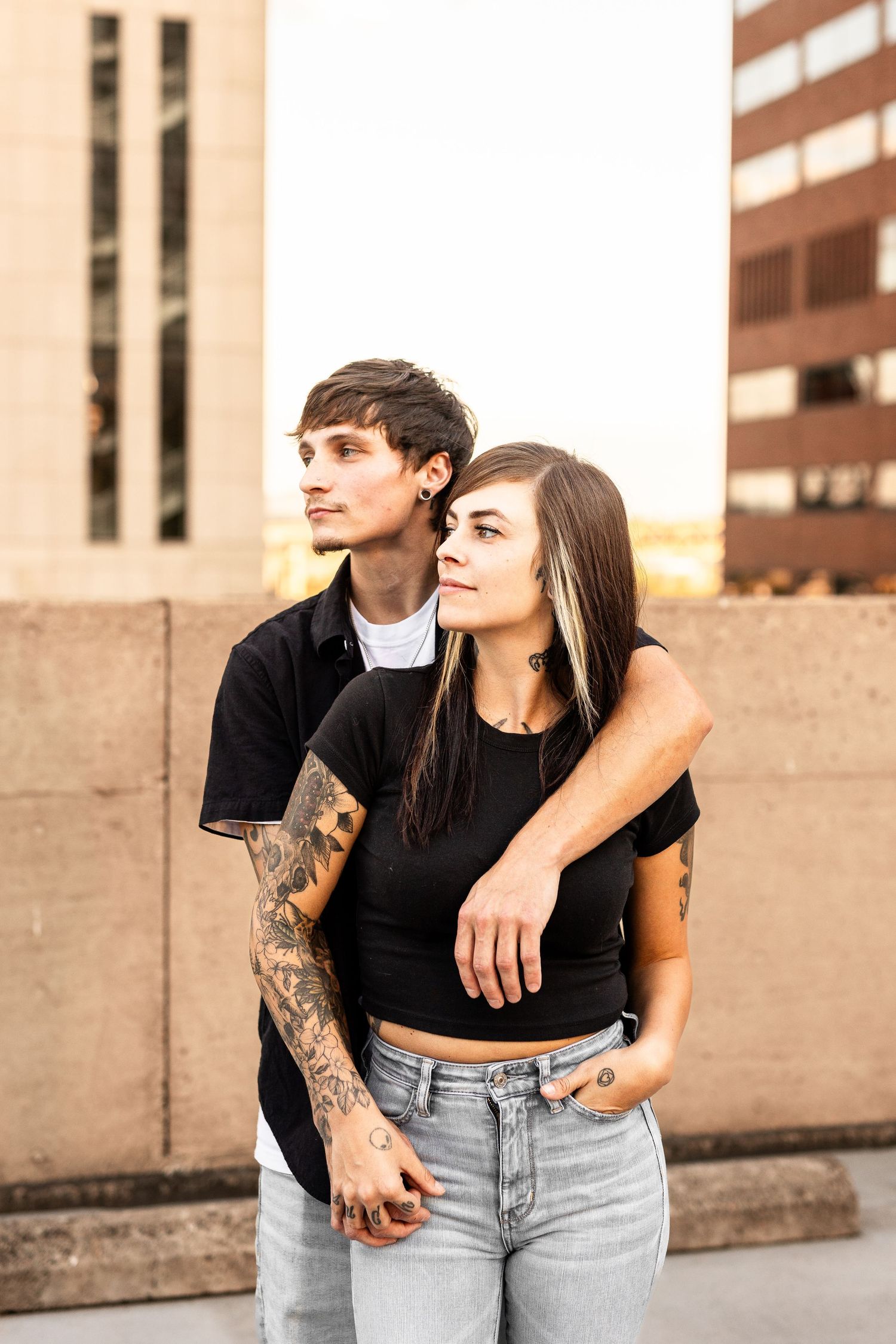A tattooed couple in black outfits embraces on a rooftop with city buildings in the background at sunset.