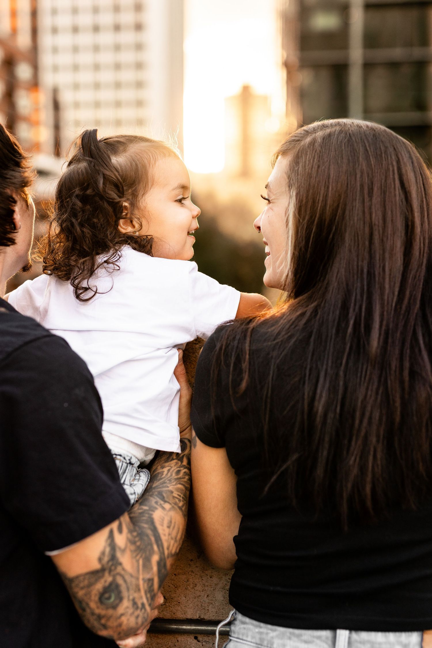 Family moment at sunset, tattooed parent holds smiling toddler while woman with dark hair laughs outdoors.