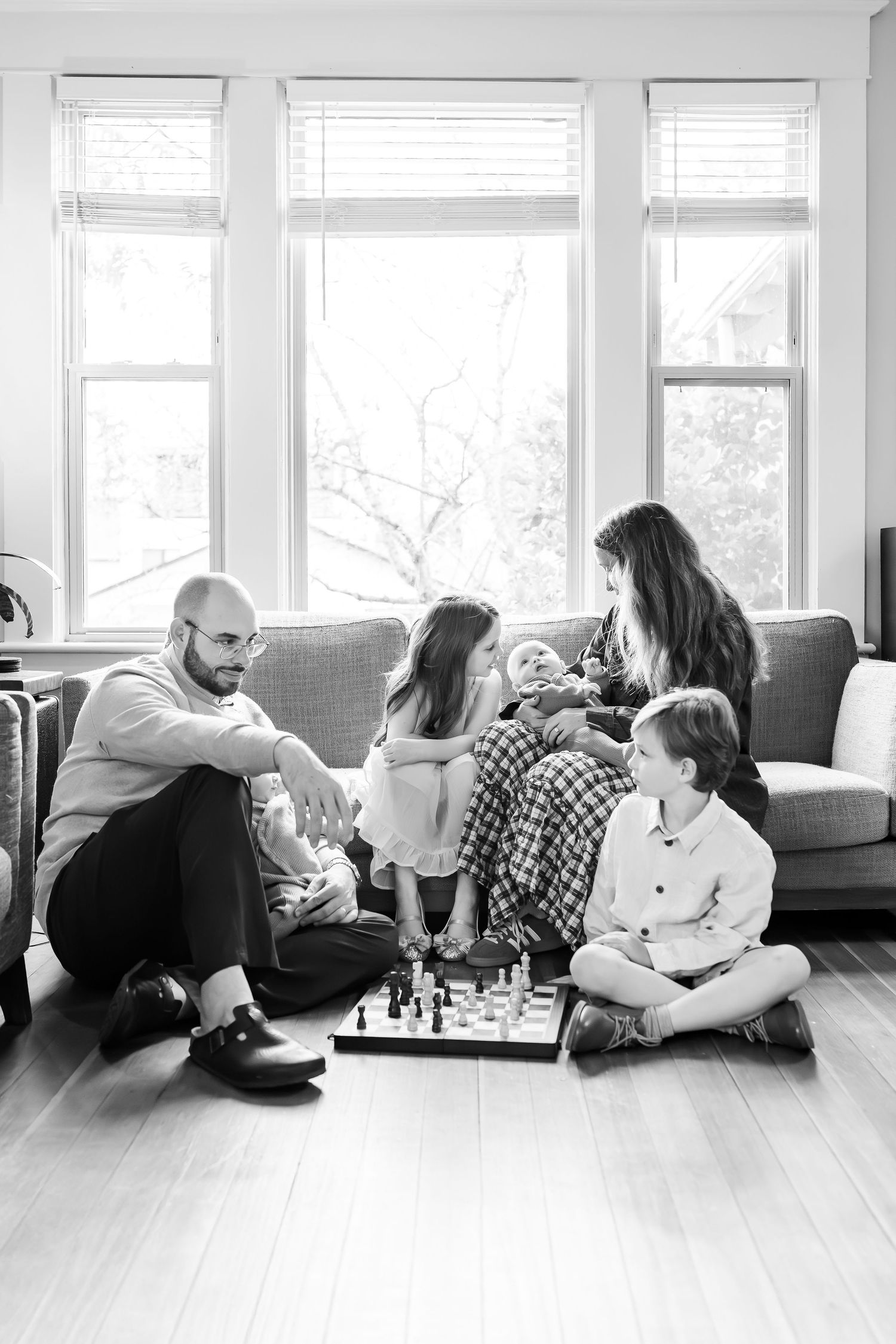 Black and white photo of a family playing chess together in a cozy living room with large windows.