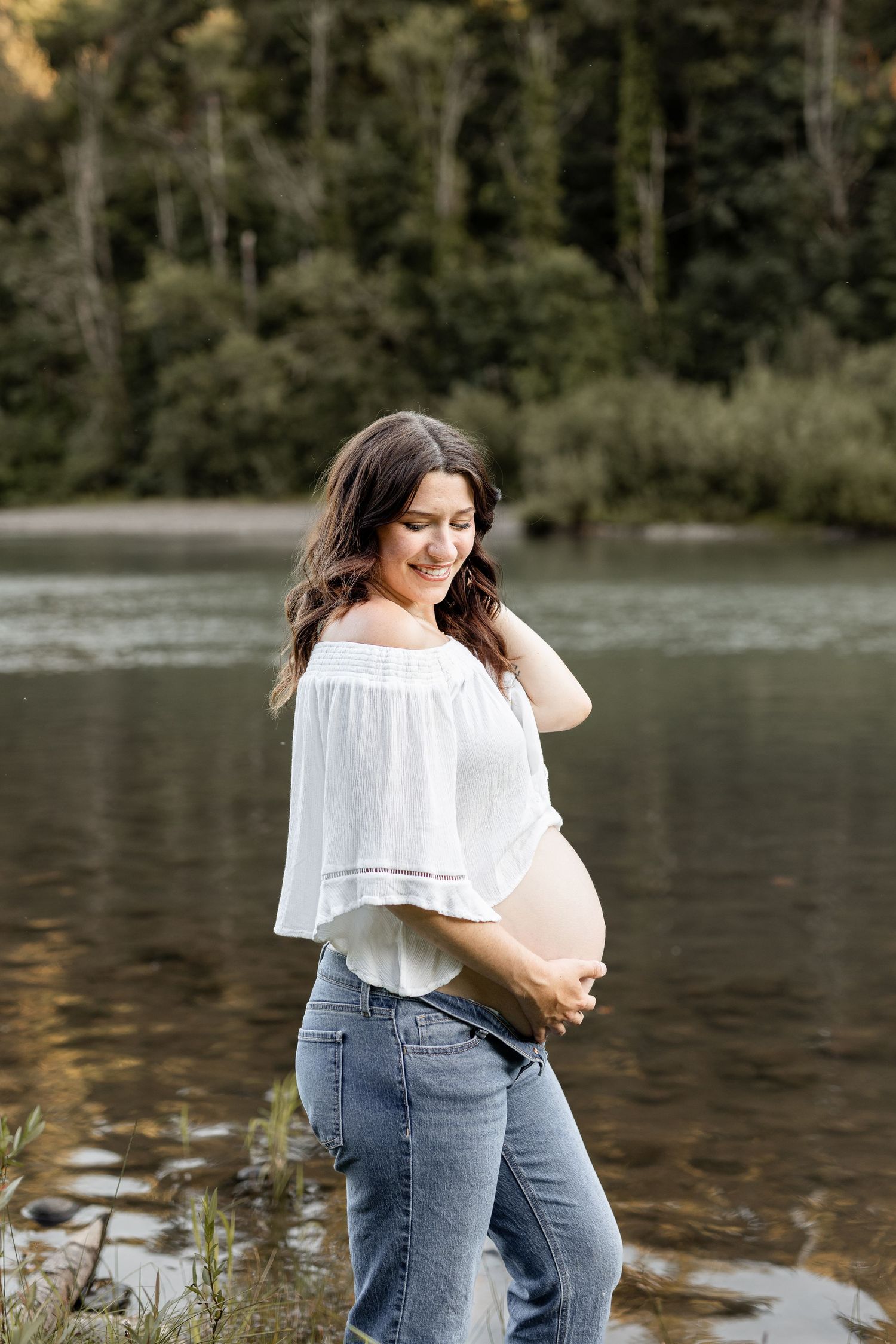 Smiling pregnant woman in white off-shoulder top and jeans cradling her baby bump near a serene forest lake.