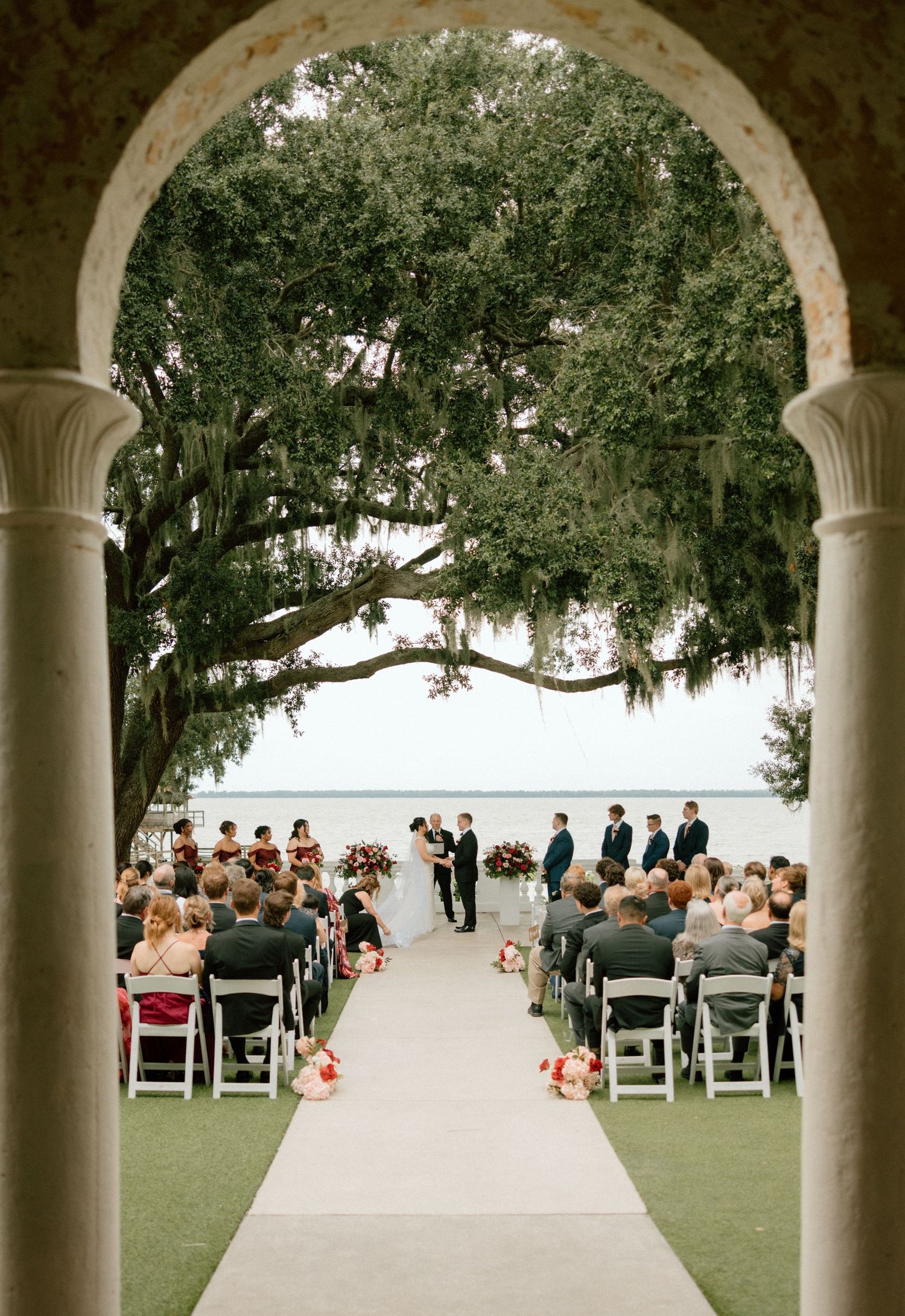 Wedding ceremony setup with white columns framing a waterfront view as guests gather on chairs on the lawn.