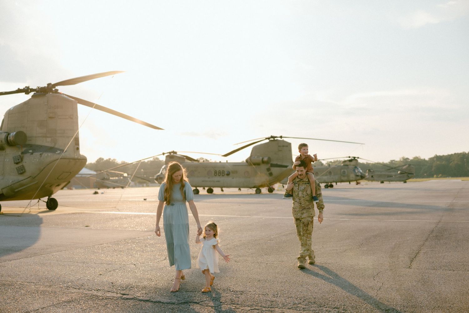Military family walking together on airfield tarmac with helicopters in background during sunset.