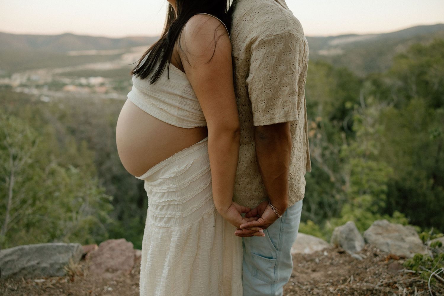 A couple embraces on a hillside overlooking a valley, with the person wearing a flowing white maternity dress.