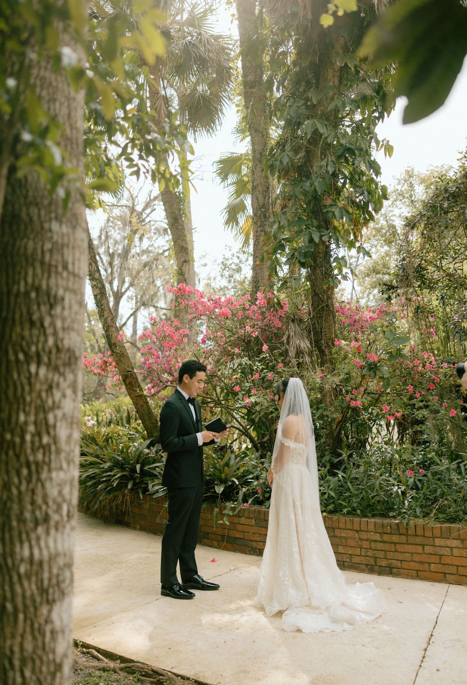 Elegant garden wedding scene with a couple on a brick-lined path surrounded by blooming pink flowers and lush trees.