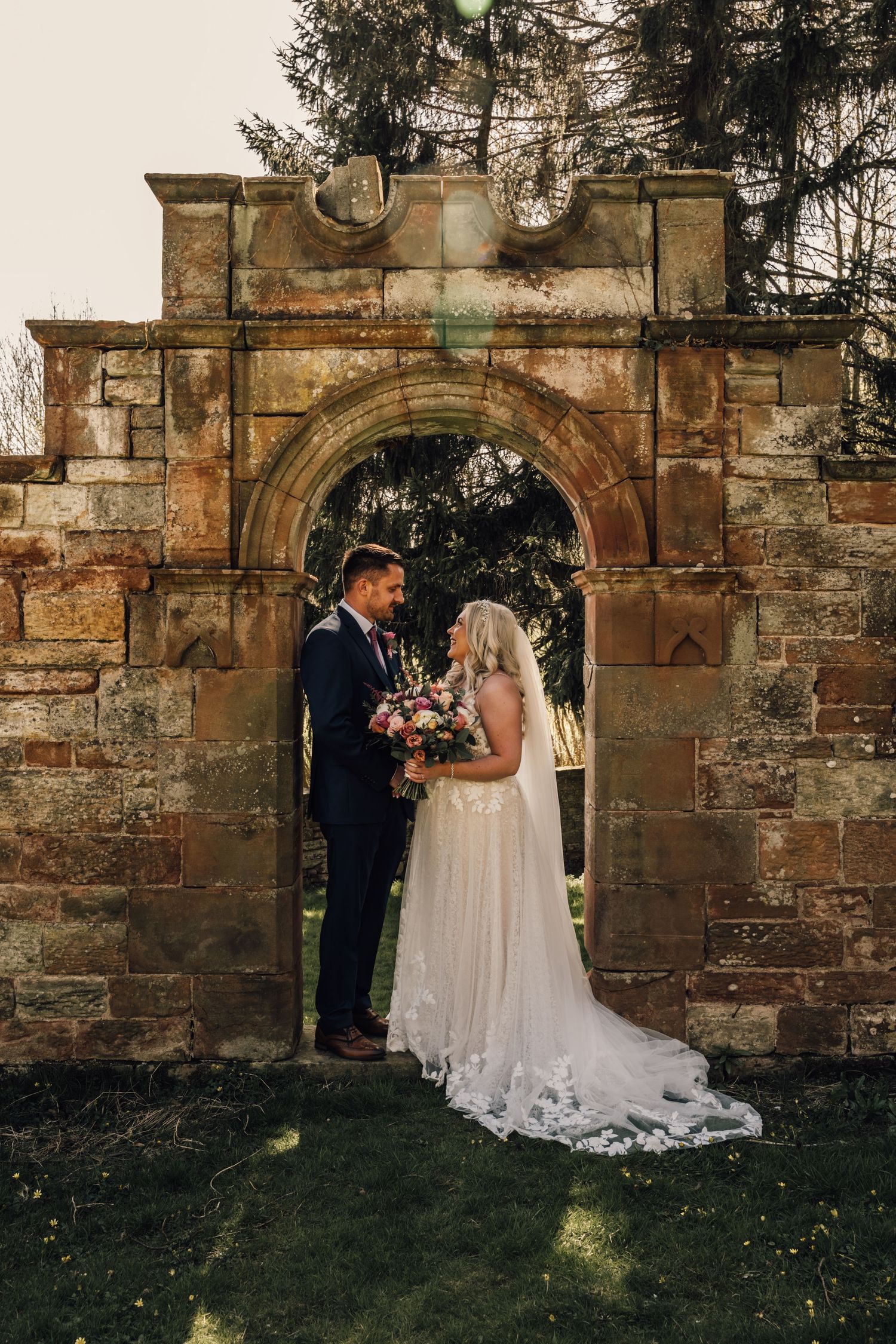 Newlyweds pose by weathered stone wall with floral arch during outdoor ceremony.