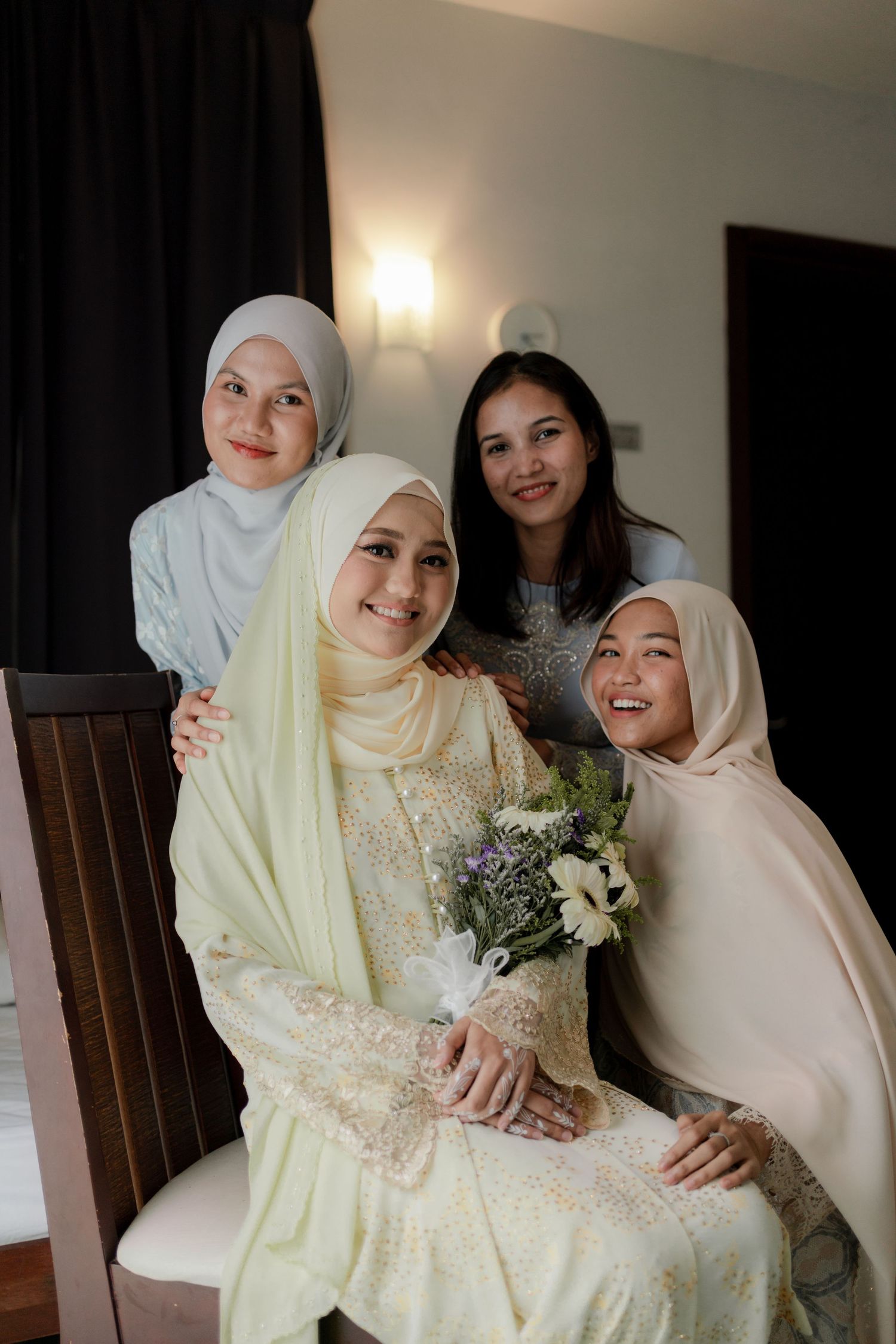 A bride in a light yellow traditional dress and hijab poses with loved ones during a wedding celebration.
