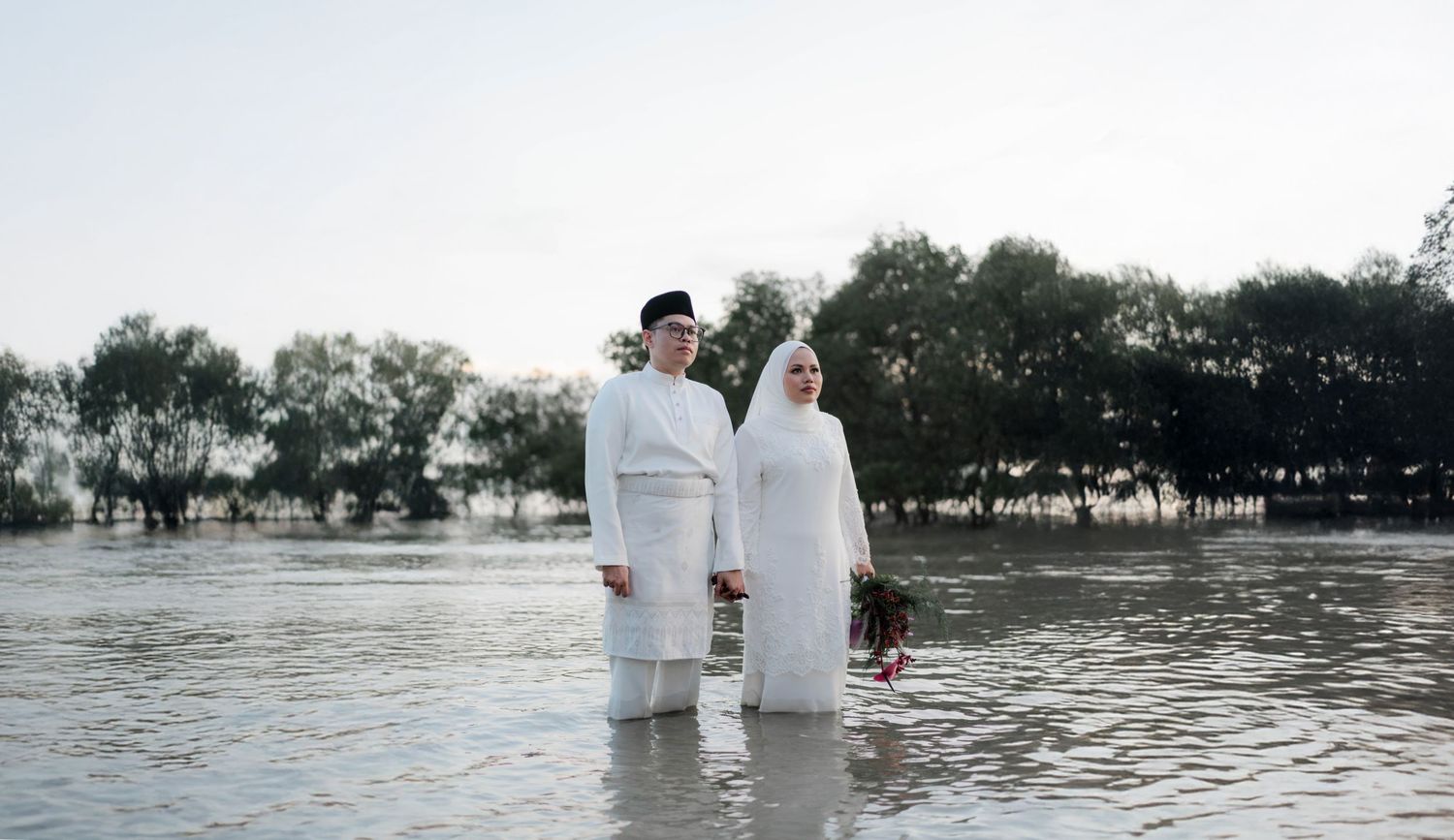 Couple in white traditional Malay attire standing in shallow floodwaters surrounded by trees.