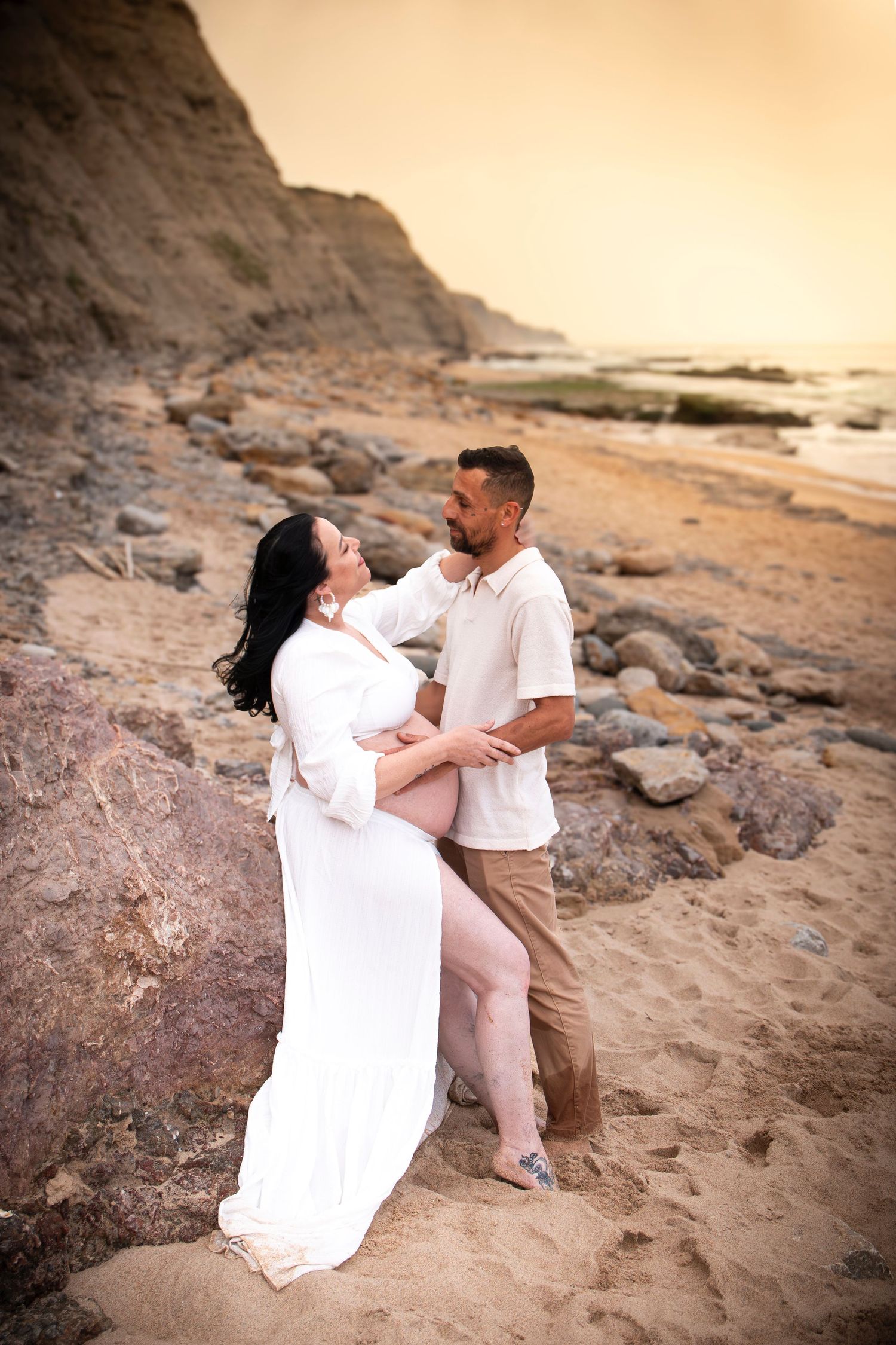 Pregnant couple embracing on a sandy beach at sunset, woman in white dress, man in beige outfit, rocky cliffs behind.