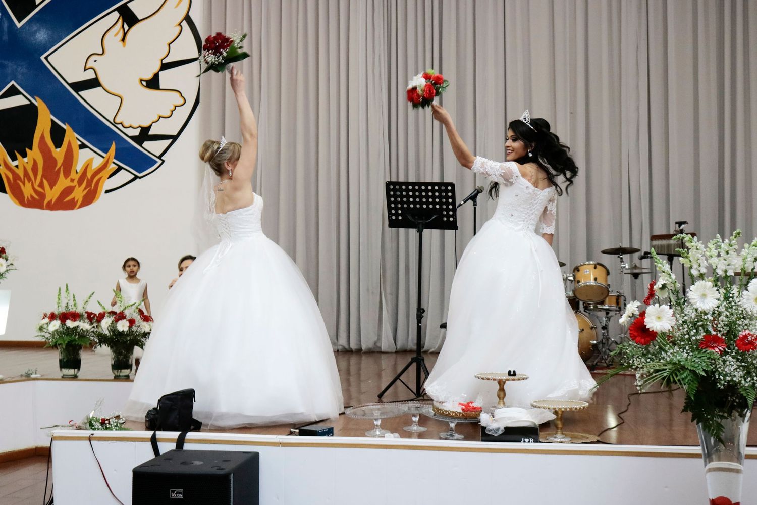 Two brides in white ballgowns perform a dance routine on stage during their wedding celebration.