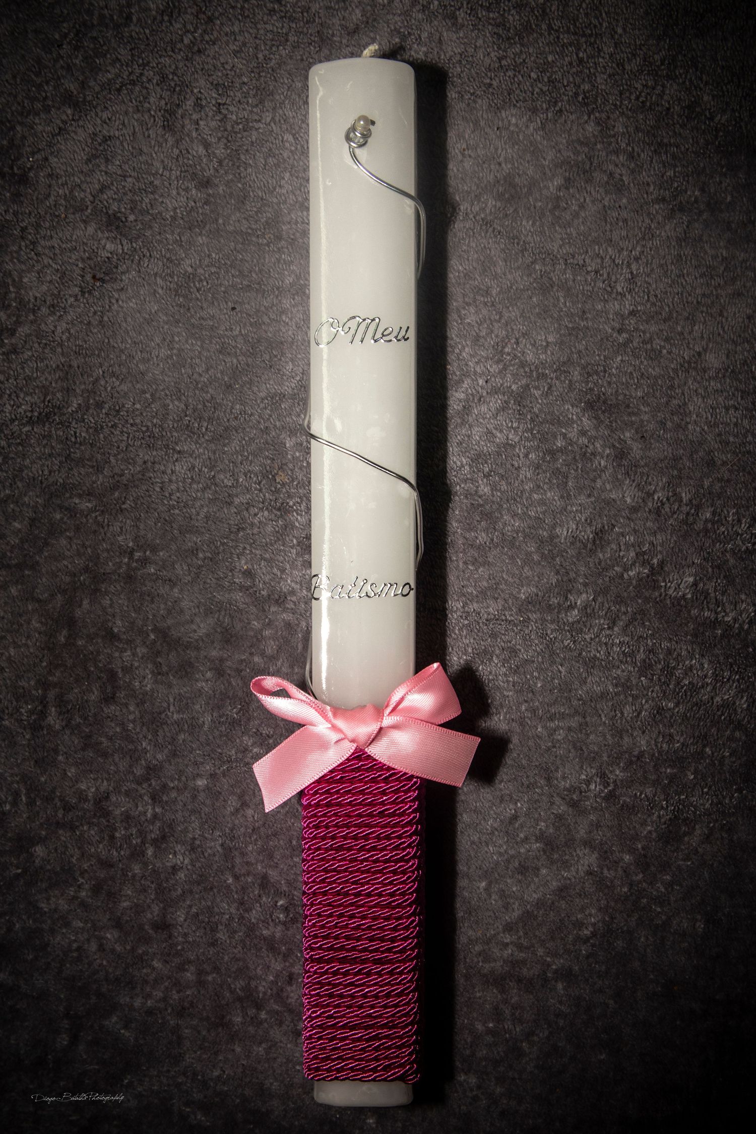 White ceremonial candle adorned with burgundy fabric wrap and pink bow on dark textured background.
