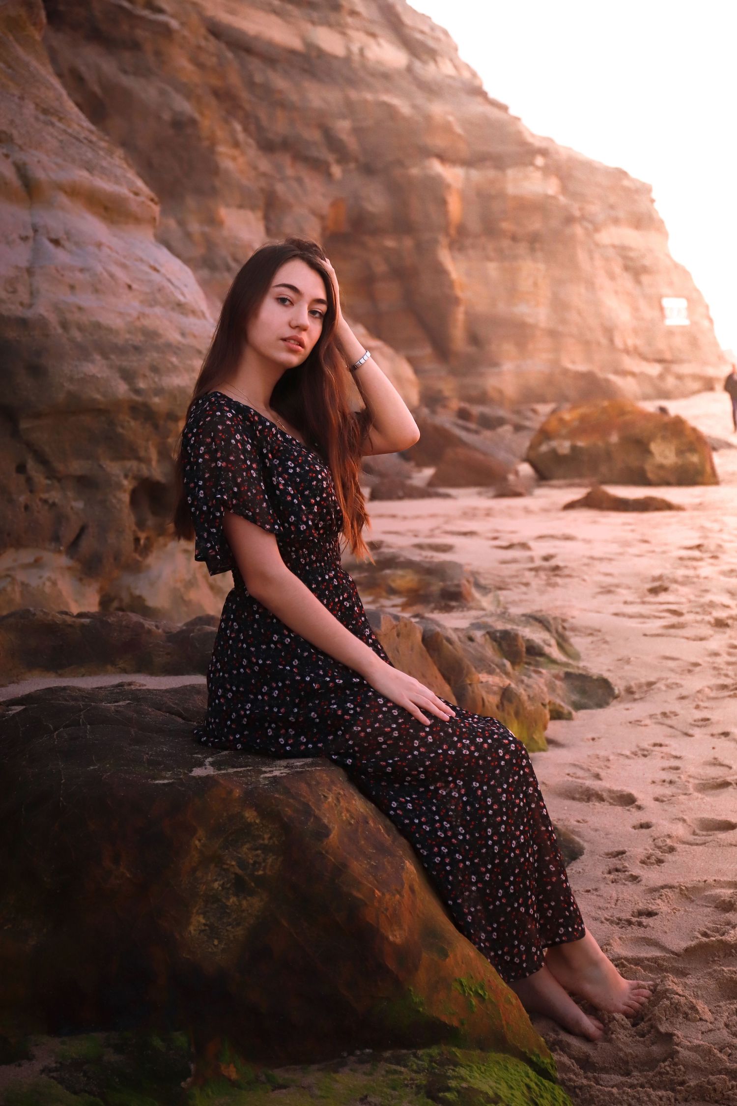 A woman in a polka dot dress sits thoughtfully on coastal rocks during golden hour at sunset.