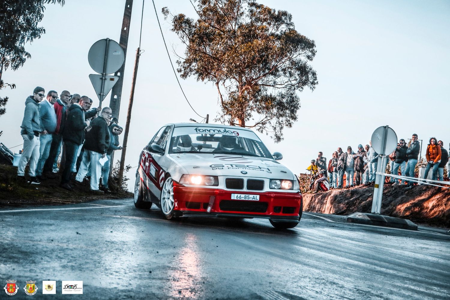 Red BMW rally car cornering at speed with brake lights illuminated and spectators watching.
