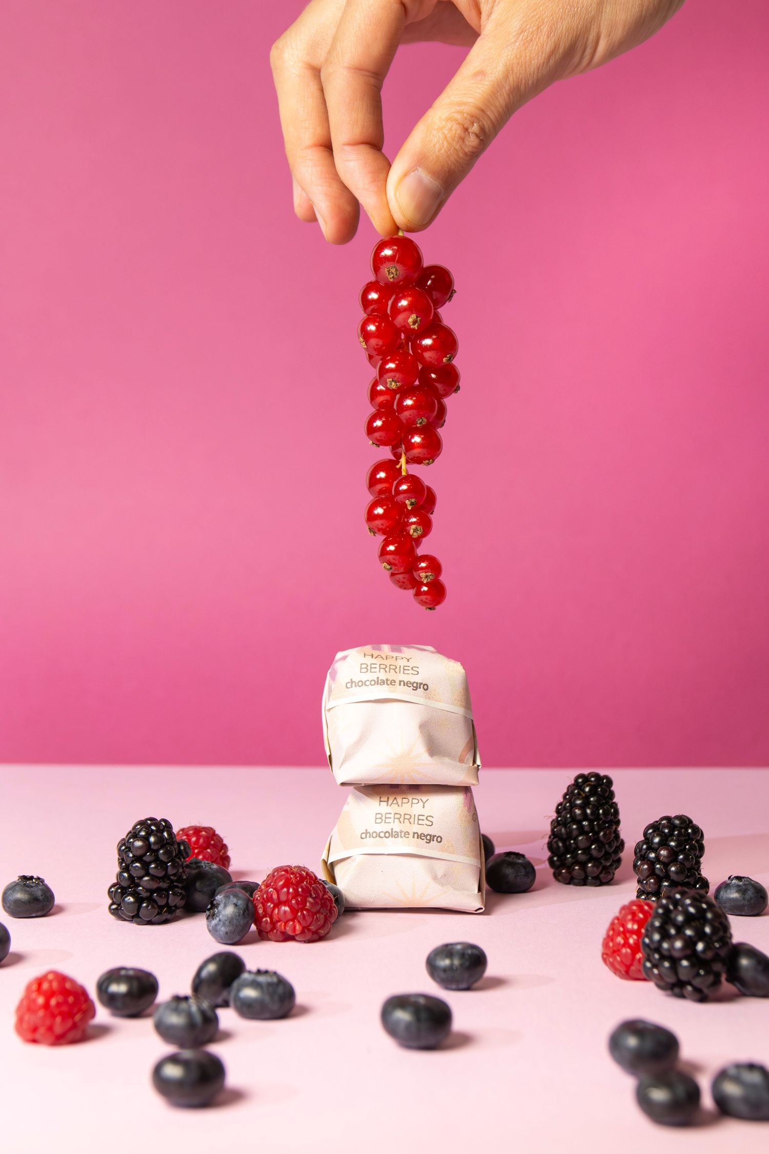 Fresh red currants being held over beauty product containers with scattered berries on pink background.