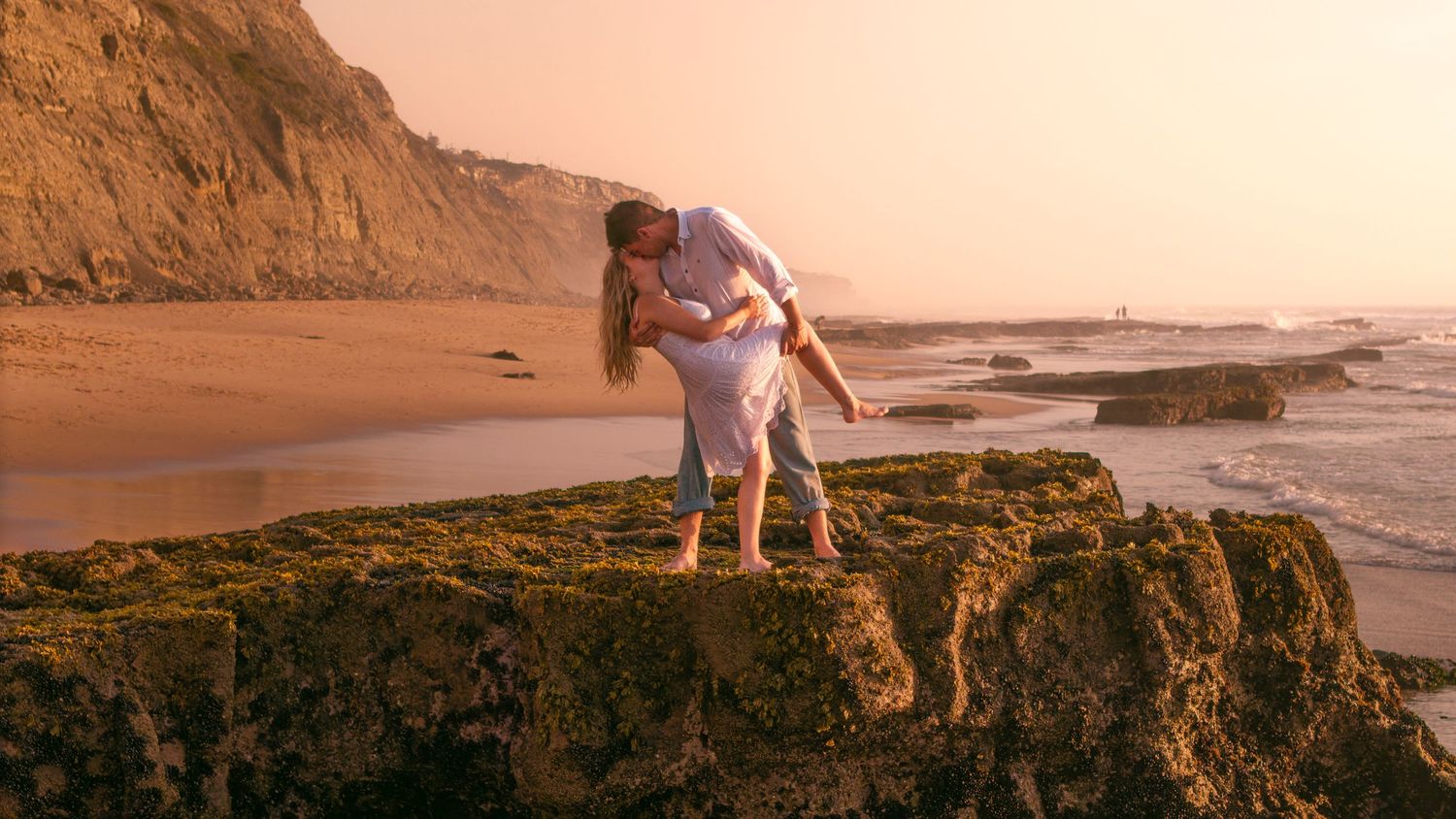 A romantic couple shares a tender moment on scenic cliffs overlooking a beautiful sunset beach in California.
