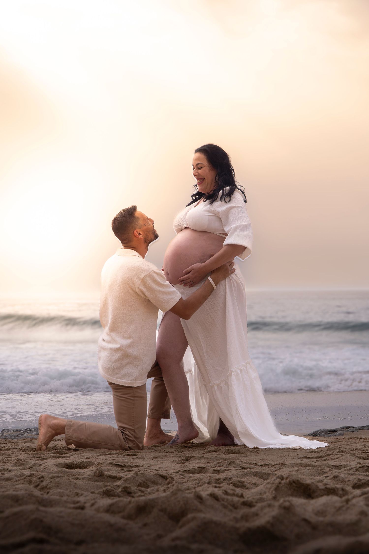 Couple on beach at sunset, man kneeling touching pregnant woman's belly, both smiling, wearing white outfits.