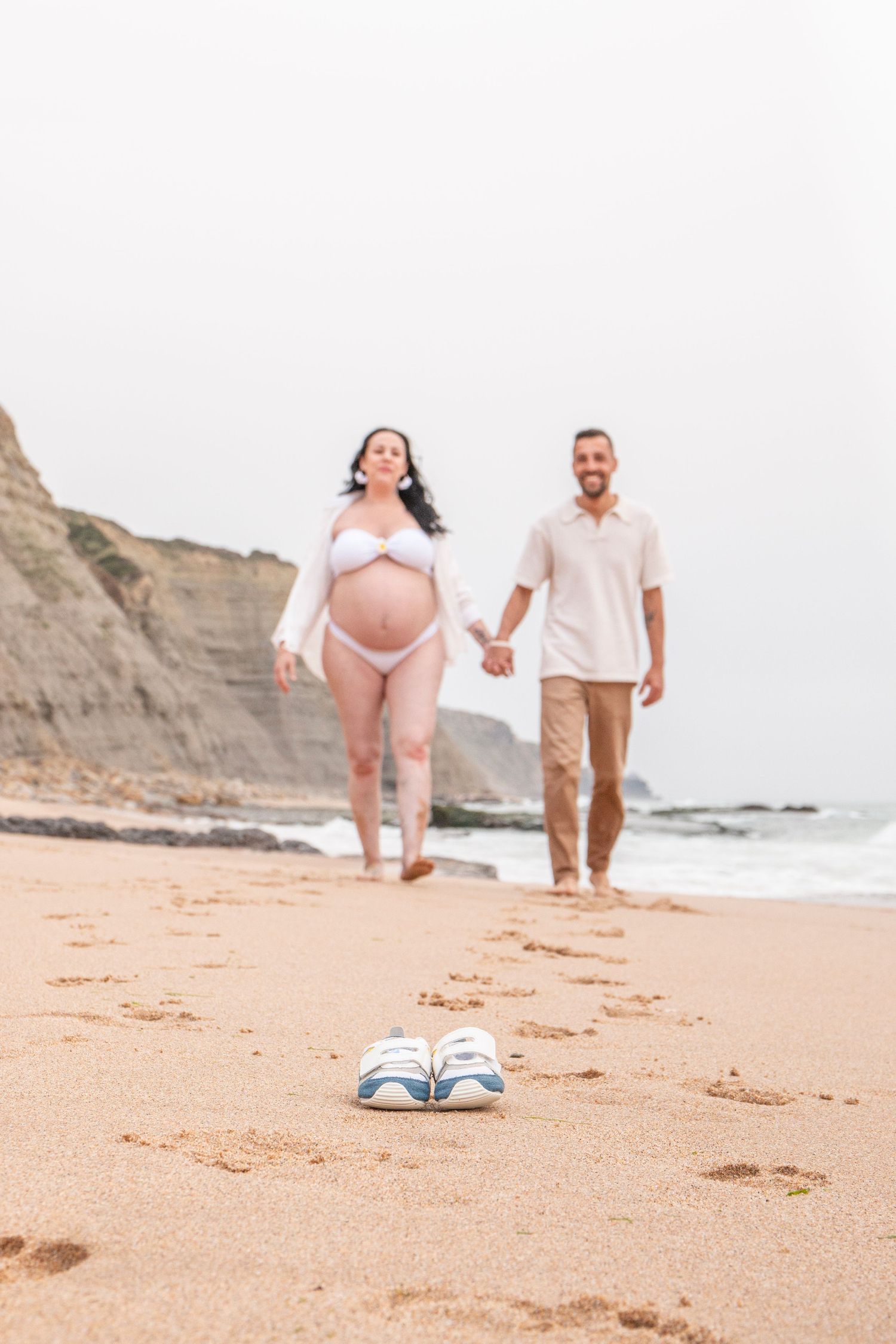Pregnant couple holding hands walking on beach with baby shoes in foreground, maternity photo shoot.