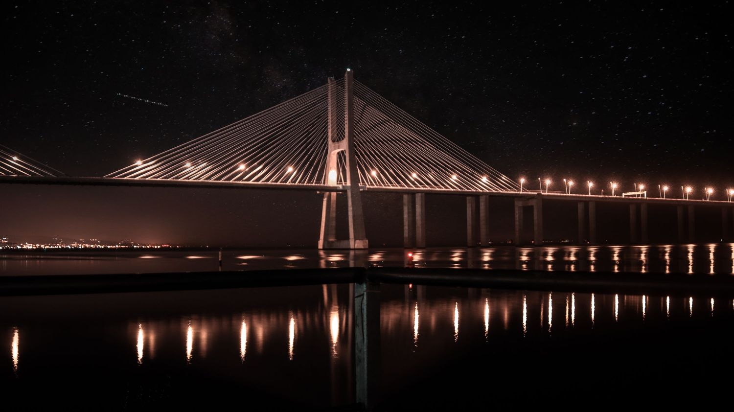 Illuminated cable-stayed bridge reflects in calm waters at night with golden lights along its span.