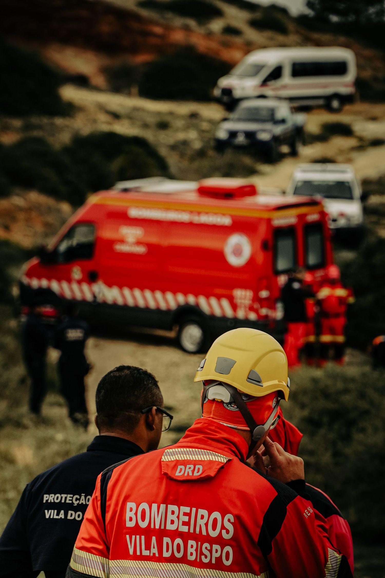 Portuguese firefighters in red uniforms from Vila do Bispo stand near emergency vehicles during a response operation.