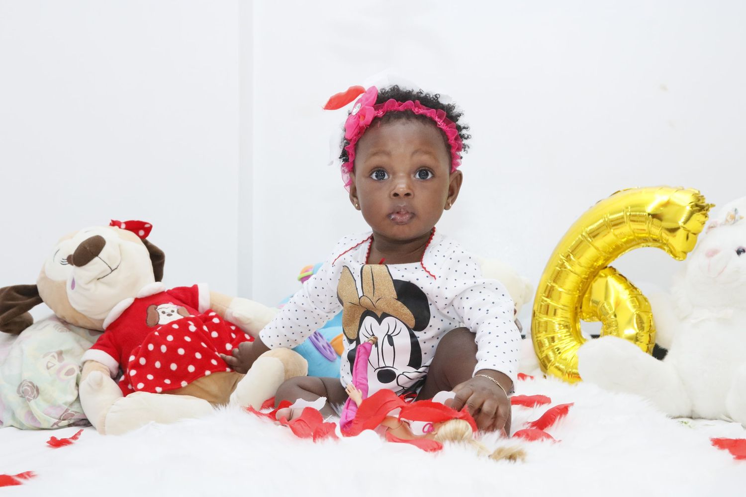 Baby in white outfit with Minnie Mouse themed decorations and toys at birthday celebration.