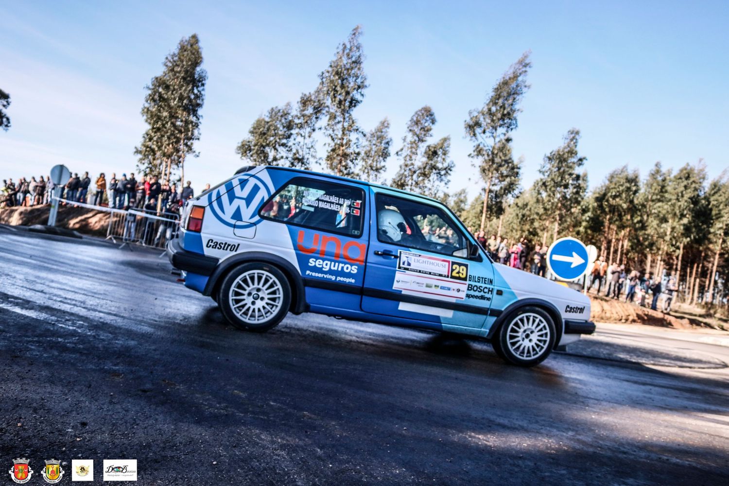 Blue rally car racing on asphalt road with spectators and trees in background.