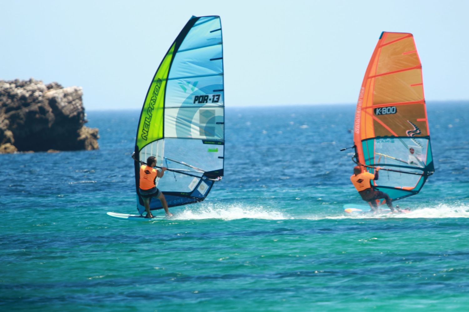 Two windsurfers racing across turquoise waters with colorful sails near rocky coast.