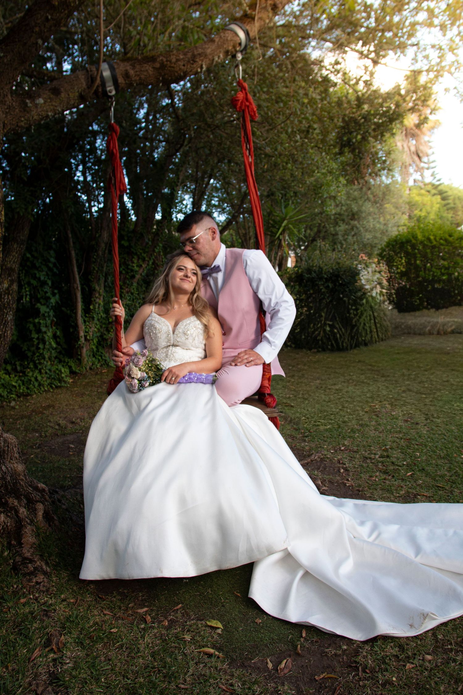 Bride in white gown sits on red rope swing as groom in pink suit leans in for a kiss outdoors.