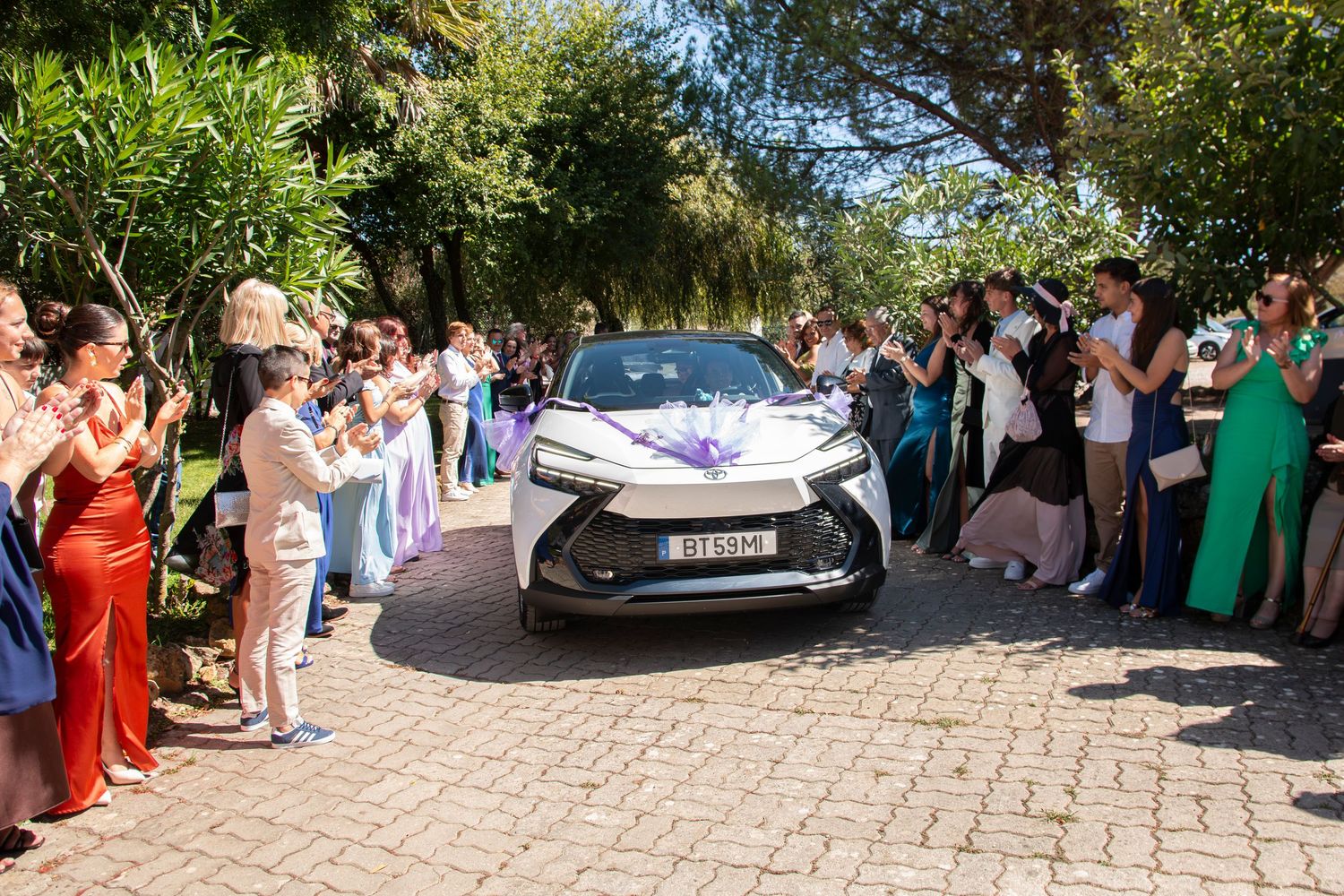 A decorated white Lexus wedding car drives through a cheering crowd at an outdoor ceremony.
