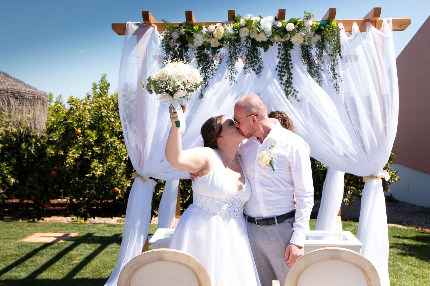 Wedding ceremony under white draped arch with flowers in outdoor garden setting.