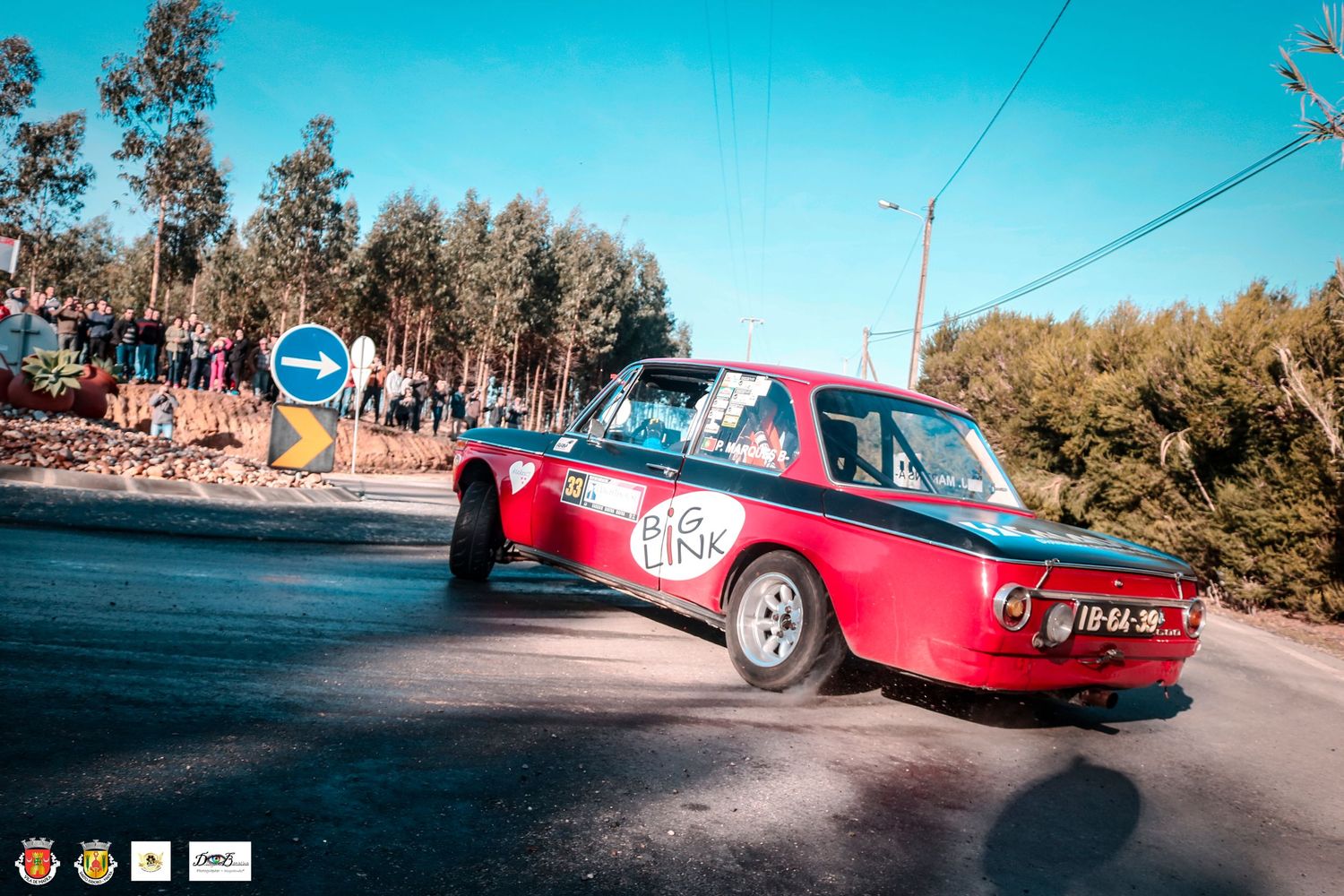 Red vintage rally car drifting through corner on asphalt road under blue sky.