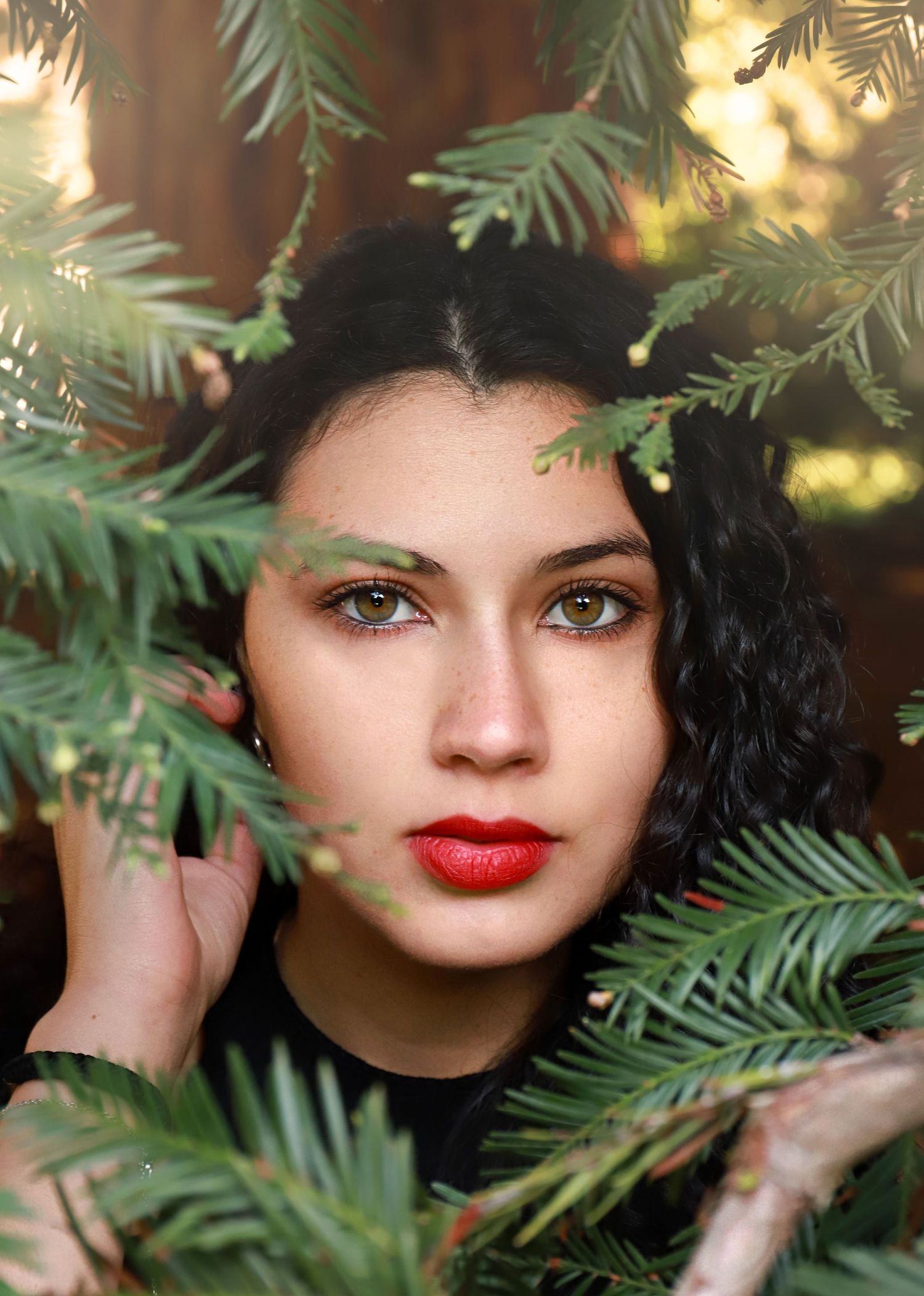 Artistic portrait through evergreen branches with red lipstick and dramatic makeup.