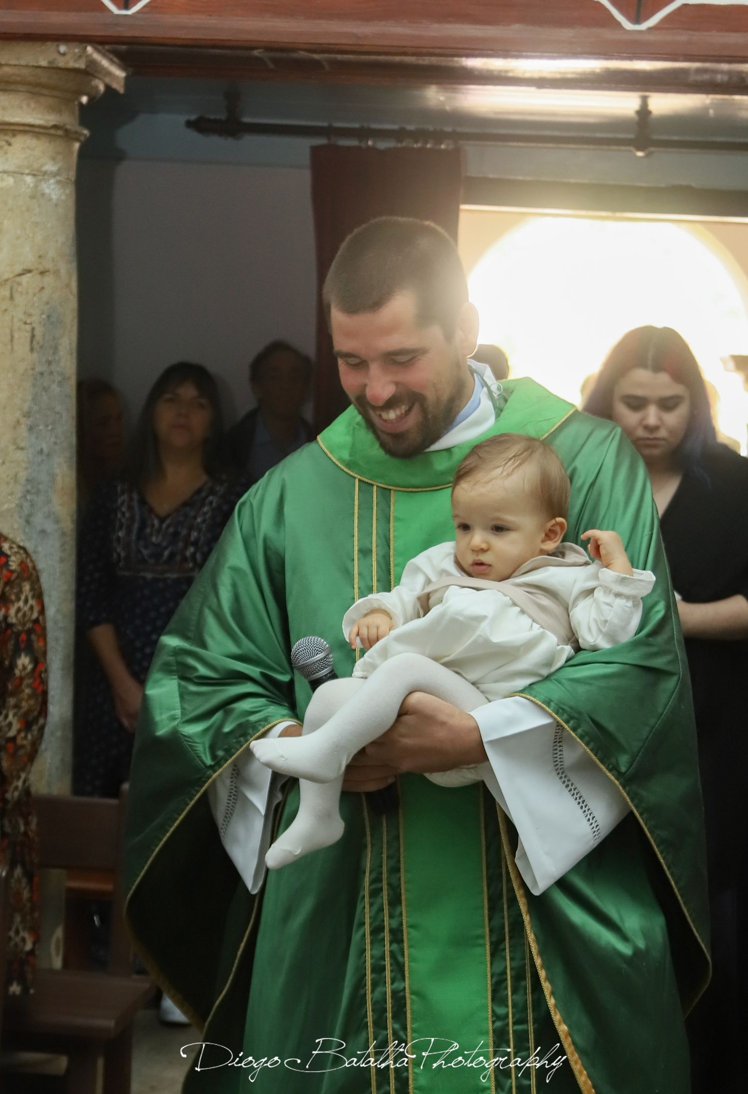 Religious ceremony with person in green robes holding baby in white gown.