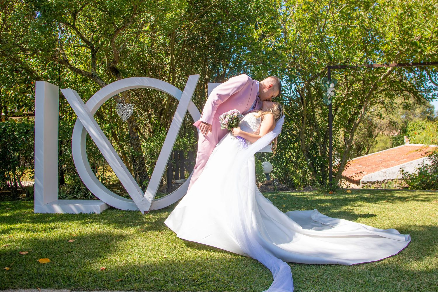 Bride and groom share a romantic dip kiss in front of a large white LOVE sign on a lush green lawn.