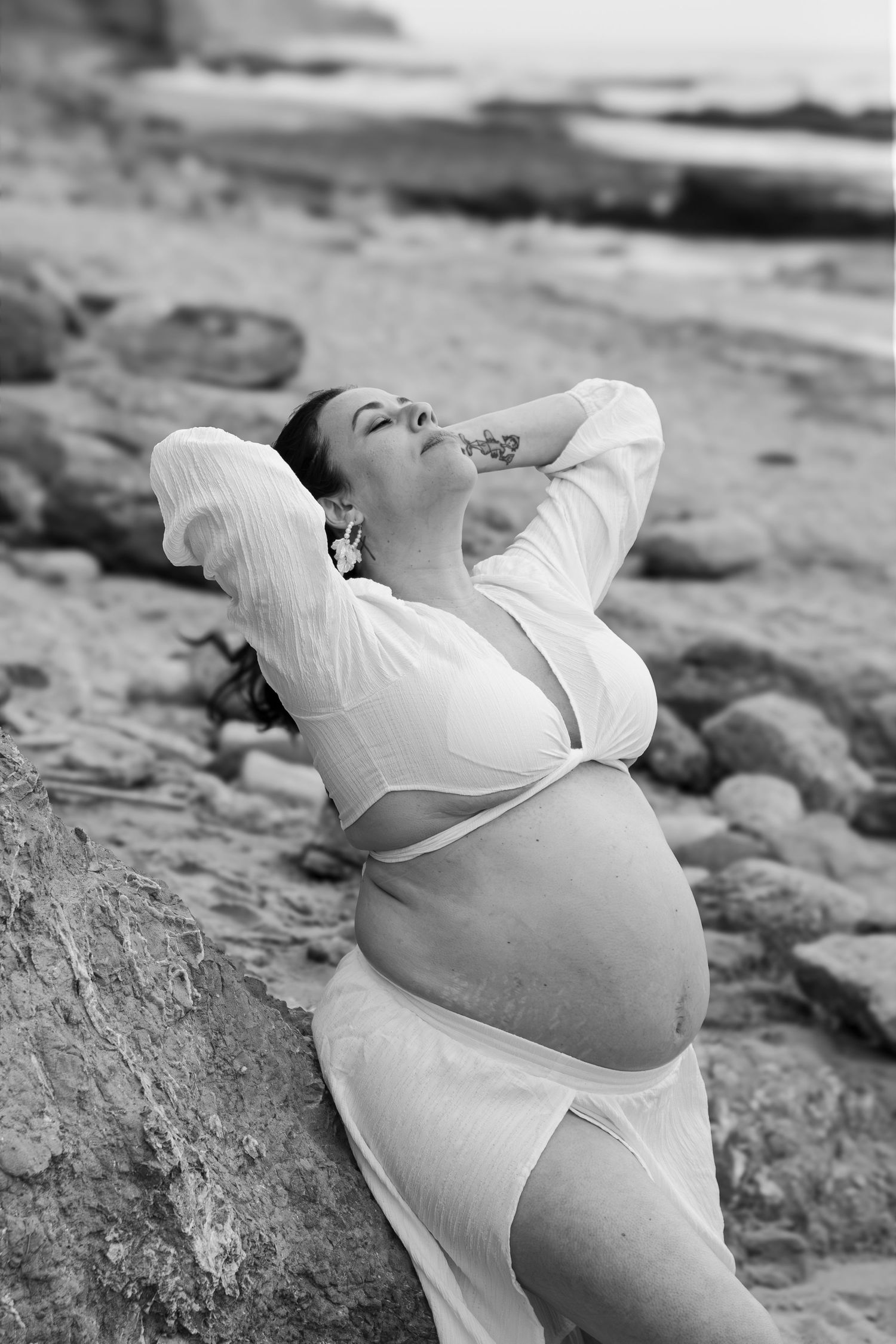 Black and white photo of a pregnant woman in a white dress posing on a rocky beach with her hands behind her head.