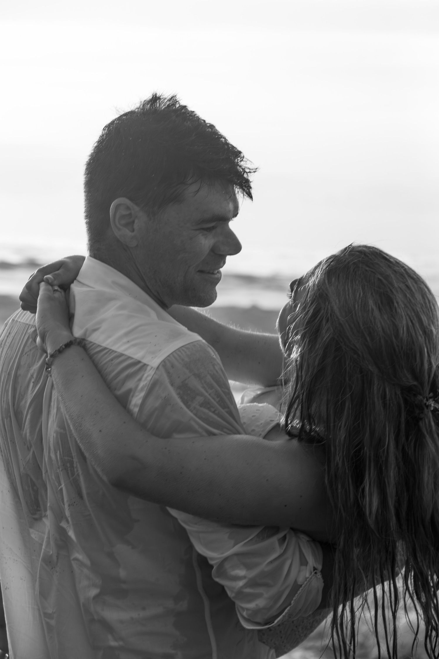 A couple embracing on the beach in black and white, sharing an intimate moment as ocean waves roll in behind them.