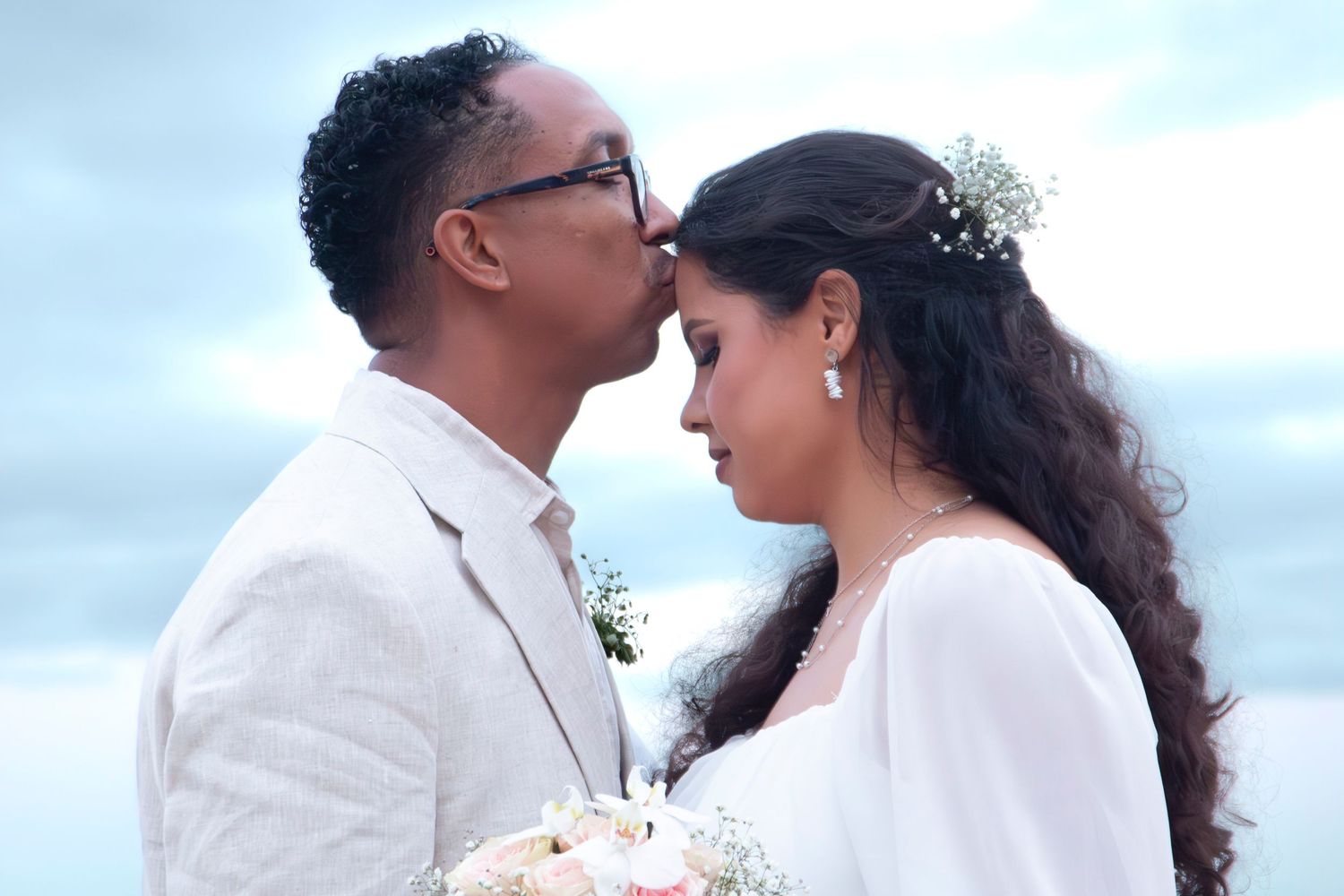 A couple in white outfits share tender moments on the beach during a romantic photoshoot at sunset.