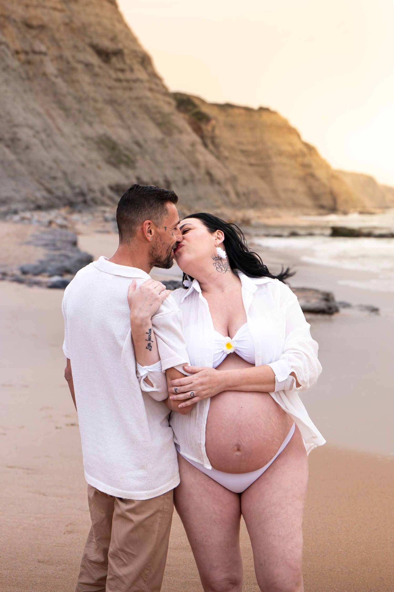 Pregnant couple sharing a kiss on a sandy beach at sunset, with rocky cliffs in the background.