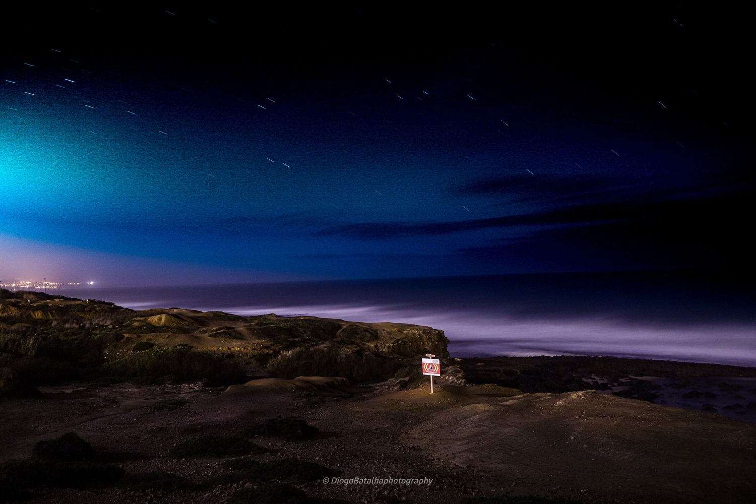A lone light illuminates a rugged coastal landscape under a dramatic blue and purple night sky with clouds.