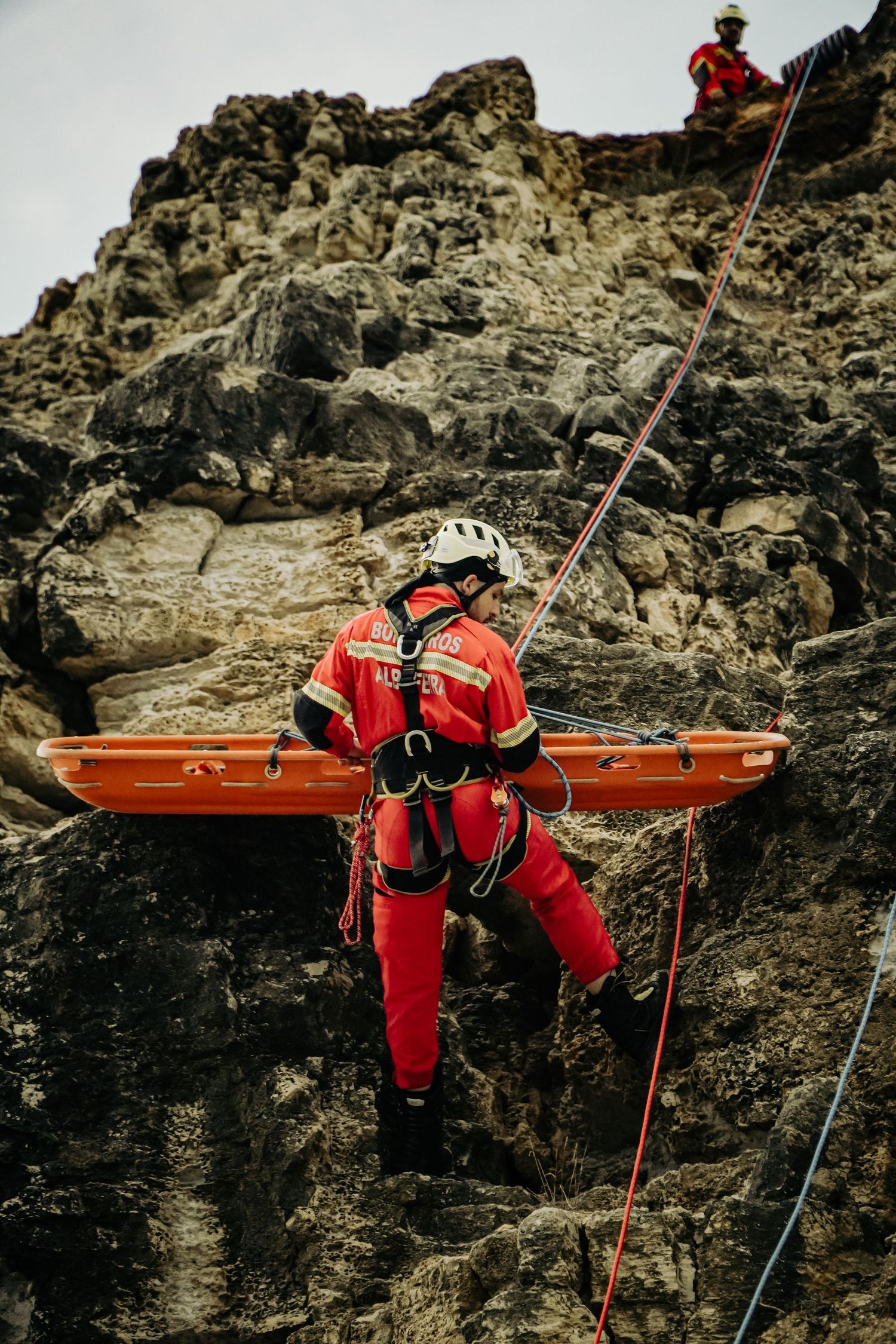 A search and rescue worker in red gear rappels down a rocky cliff face while carrying emergency equipment.