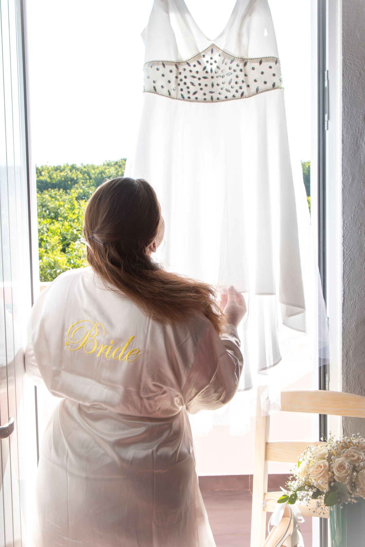 Beautiful white beaded wedding dress hanging by a window with natural sunlight.